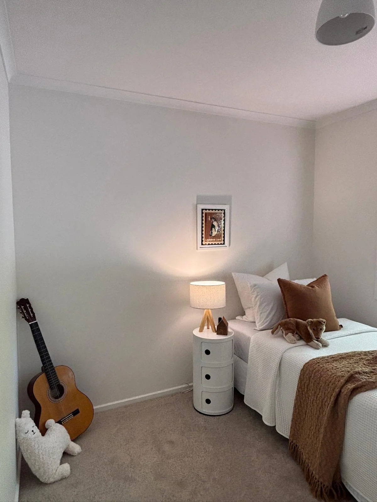 A cozy bedroom with a white bed, a white nightstand with a lamp, a teddy bear, pillows, a guitar, a stuffed animal, beige carpet, and a framed picture on the wall.