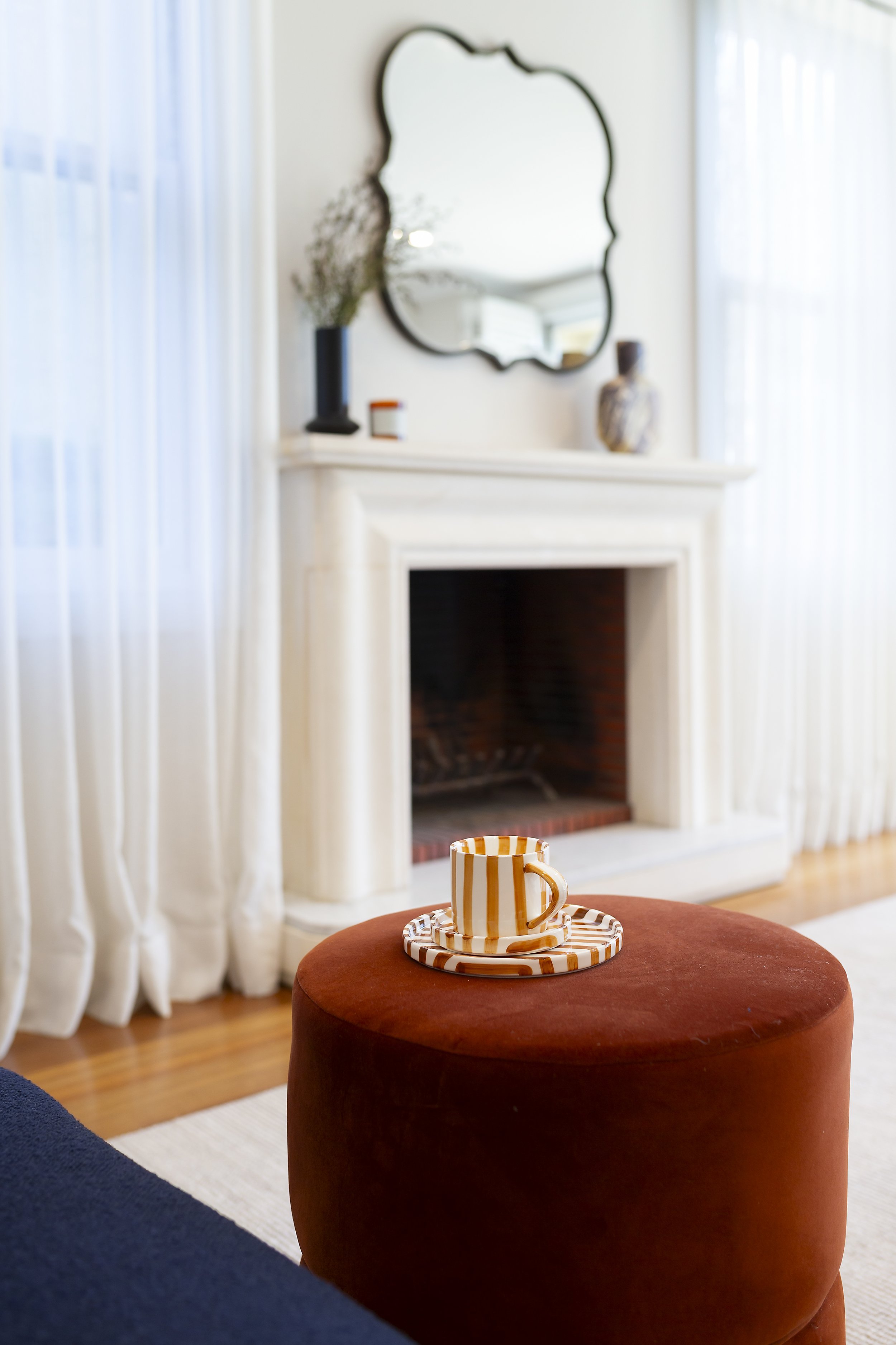 A cozy living room with a burnt orange ottoman topped with a striped coffee mug and saucer, a white fireplace, a black-framed mirror, and decorative vases on the mantel. Sheer curtains cover the windows, allowing natural light to fill the space.