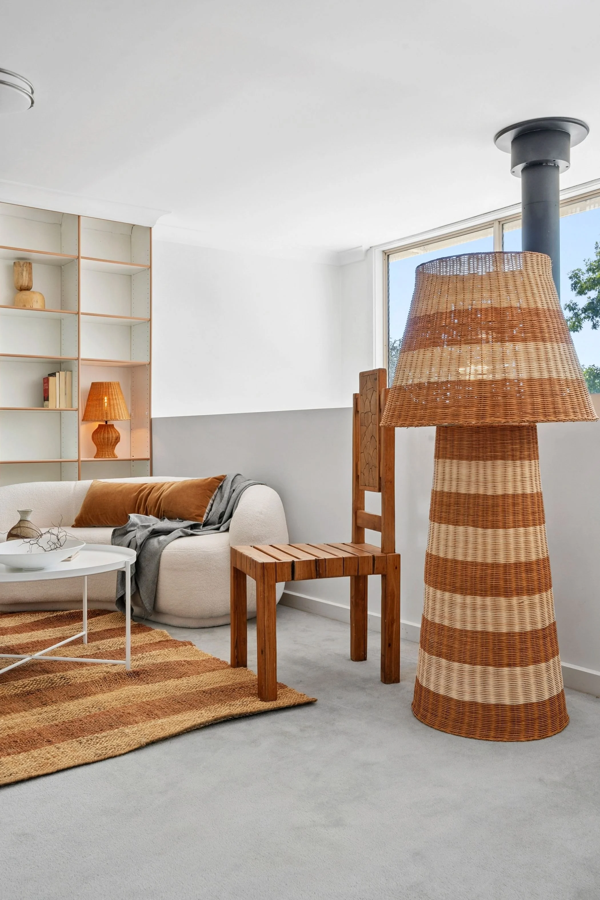 Living room with beige sofa, wooden chair, striped wicker floor lamp, white coffee table, bookcase, and a window with blue sky and trees outside.