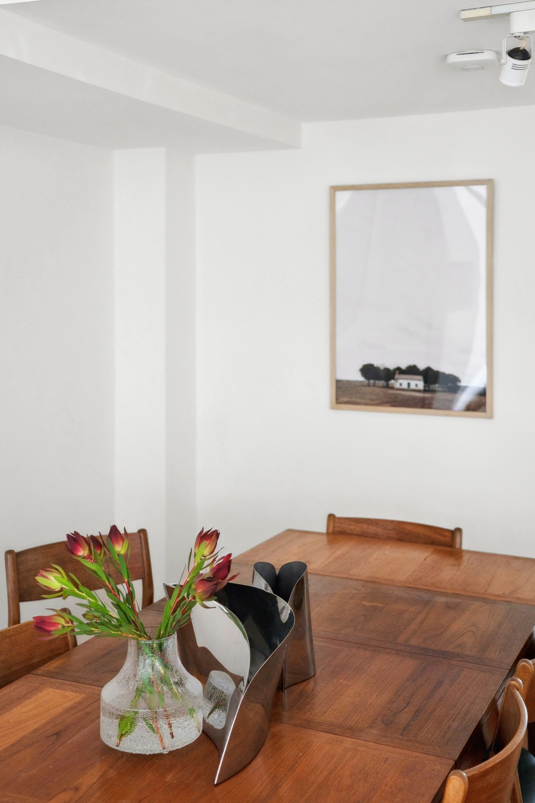 Interior of a dining room with a wooden table and chairs, a glass vase with flowers, and a framed picture on a white wall.