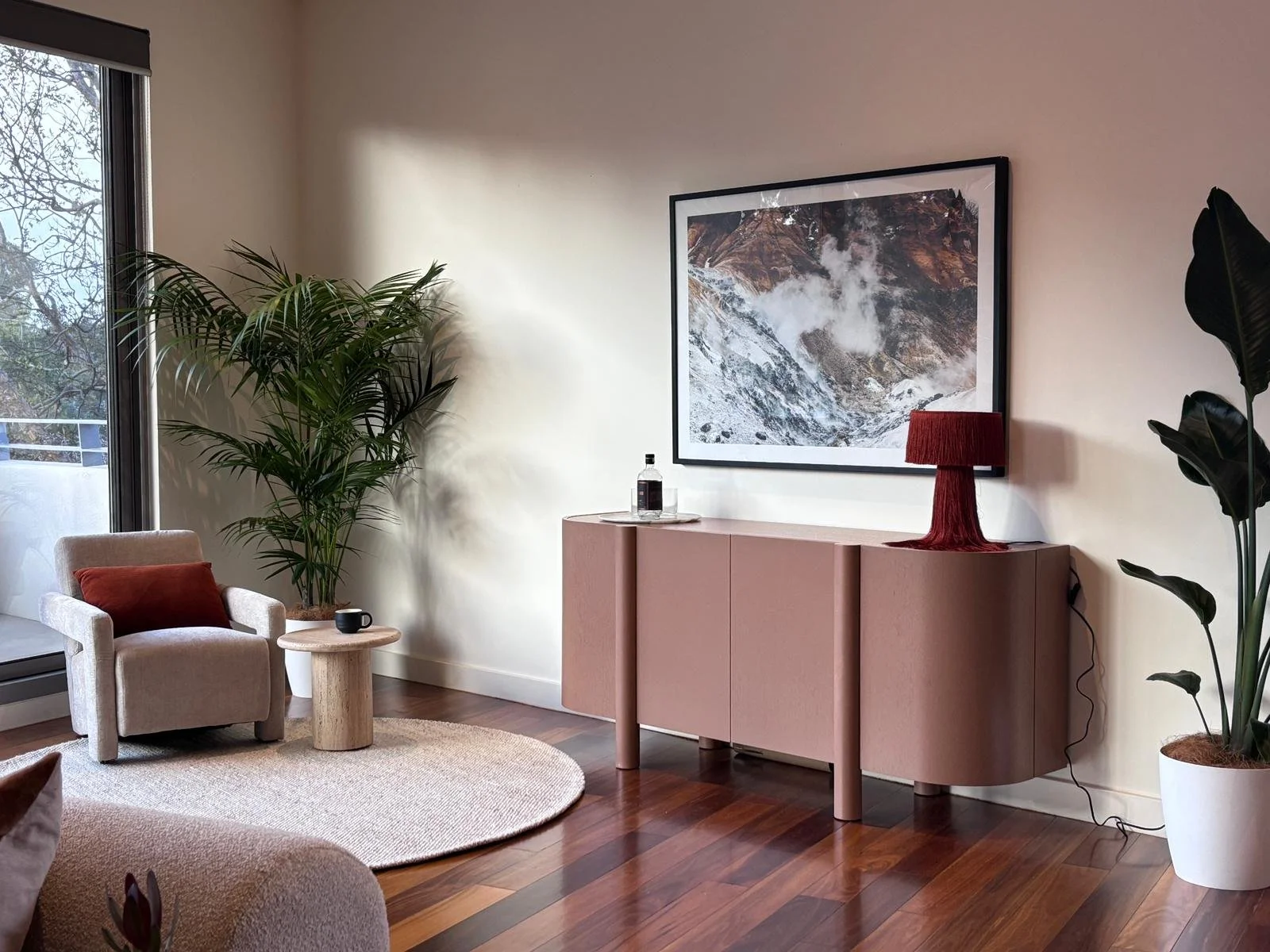 A cozy living room with a beige armchair, a small wooden side table with a mug, large green plants, a pink cabinet with a red lamp, a framed mountain landscape picture on the wall, and a sliding glass door with a view of the outdoor balcony.