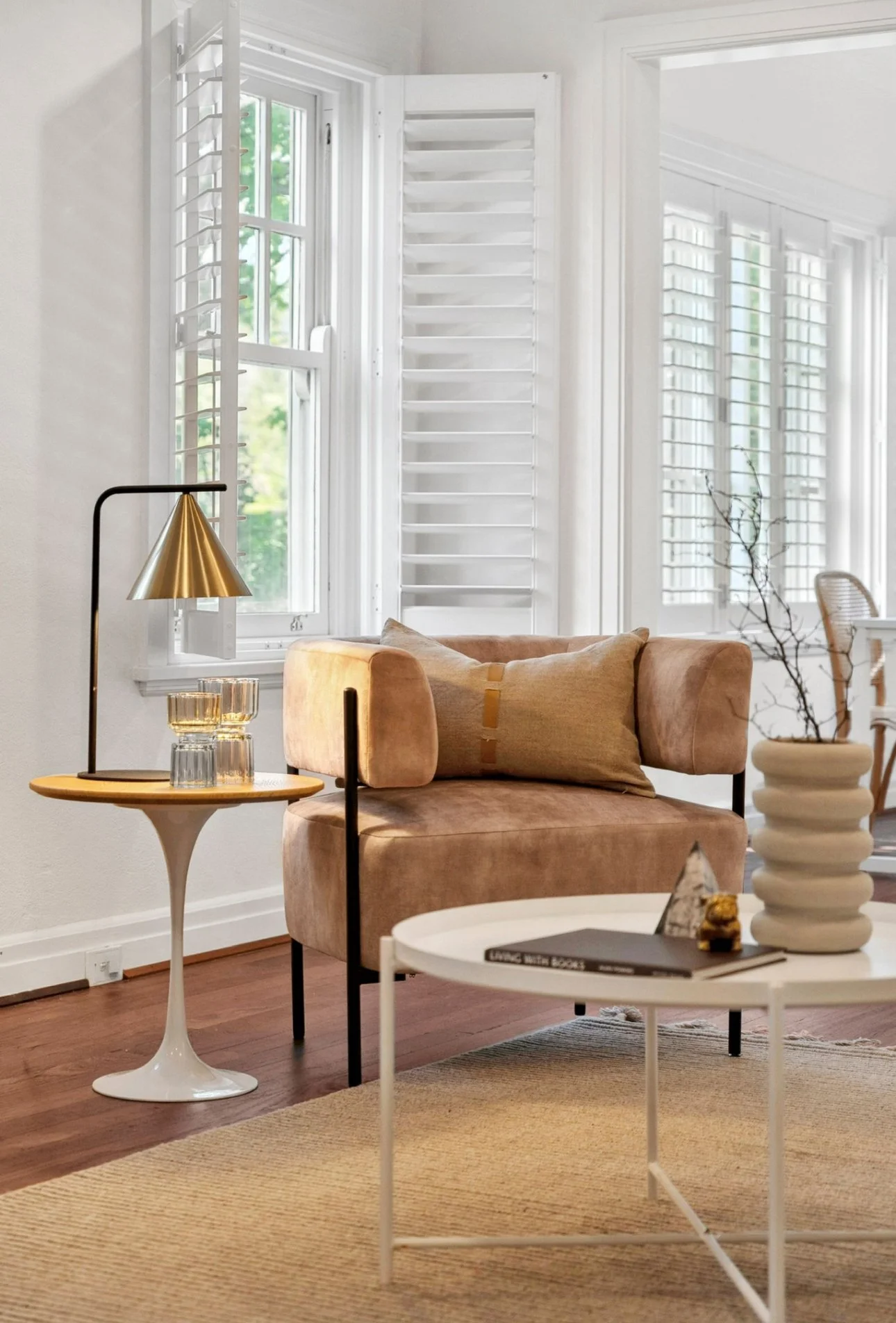 Living room with beige chairs, white round coffee table, side table with gold lamp and glasses, large windows with white shutters, decorated with a beige rug and minimal decor.