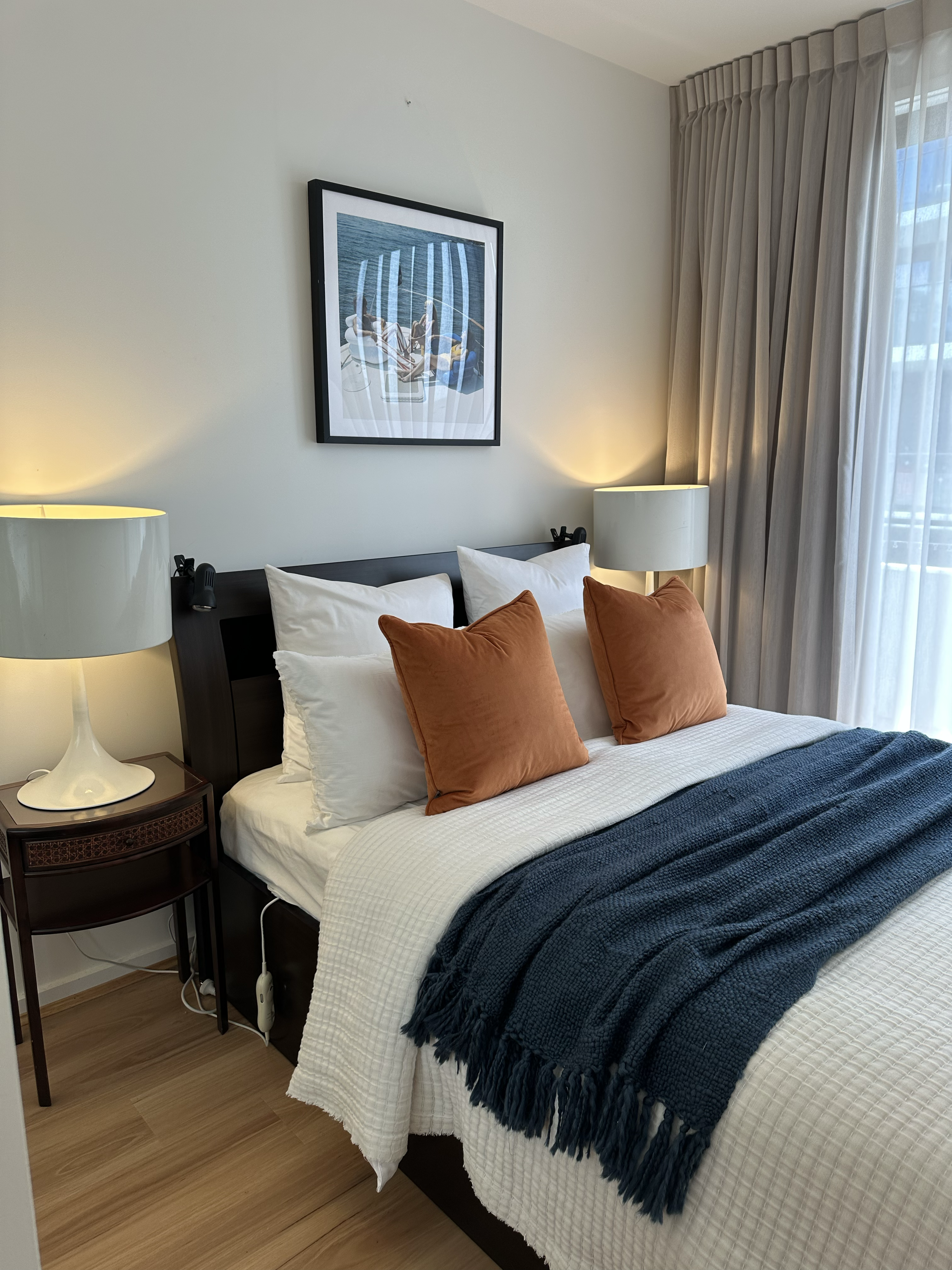 A neatly made bed with white bedding, two white pillows, two brown pillows, and a dark blue throw blanket in a hotel room. On either side of the bed are matching white lamps on dark wood nightstands. A framed art print hangs above the bed, and sheer 