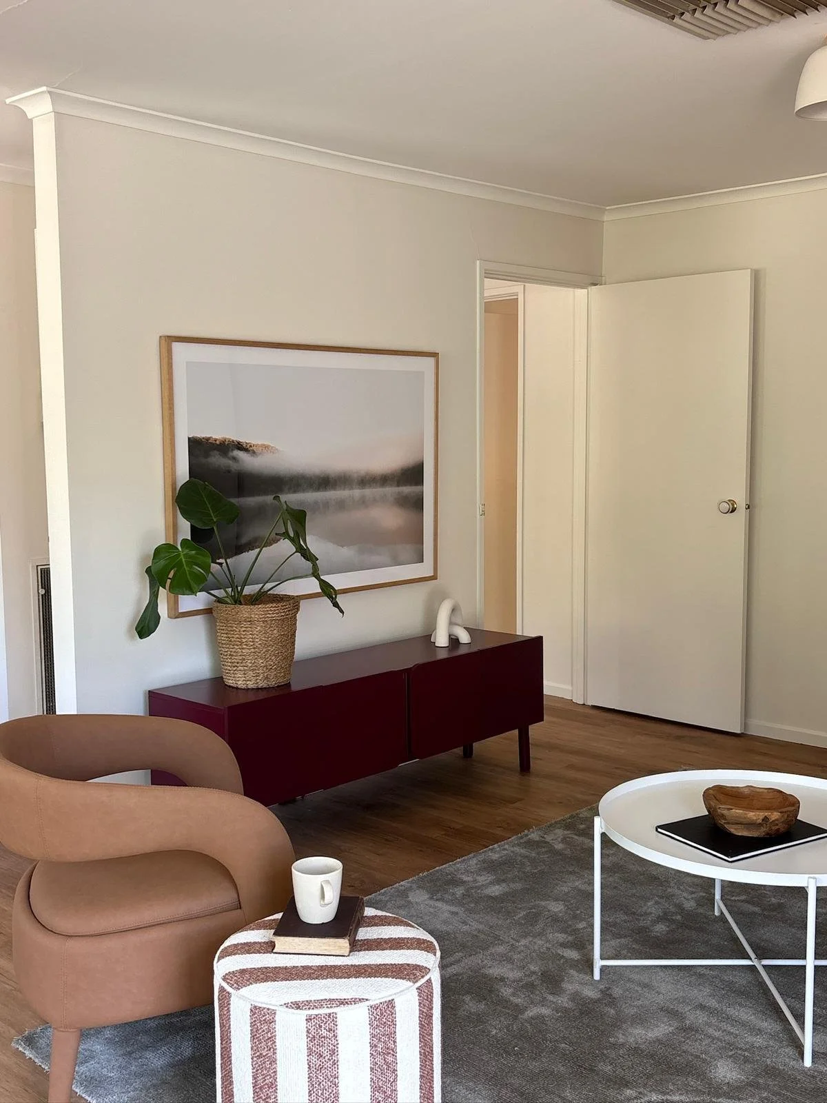 Living room with a beige armchair, a white coffee table with a wooden bowl on top, a gray rug, a red cabinet with a potted plant and decorative object, and a large framed black-and-white landscape photo on the wall.