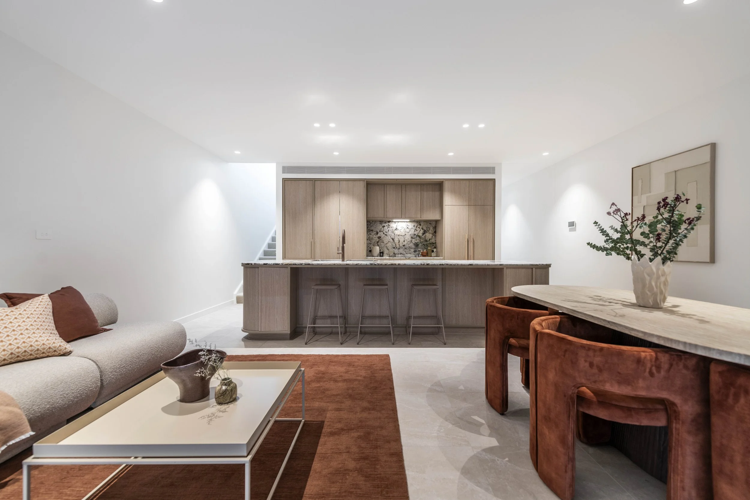 Modern living room and kitchen with beige sofa, white coffee table, orange area rug, rustic wooden dining table with rust-colored chairs, and a kitchen with light wood cabinetry and marble backsplash.