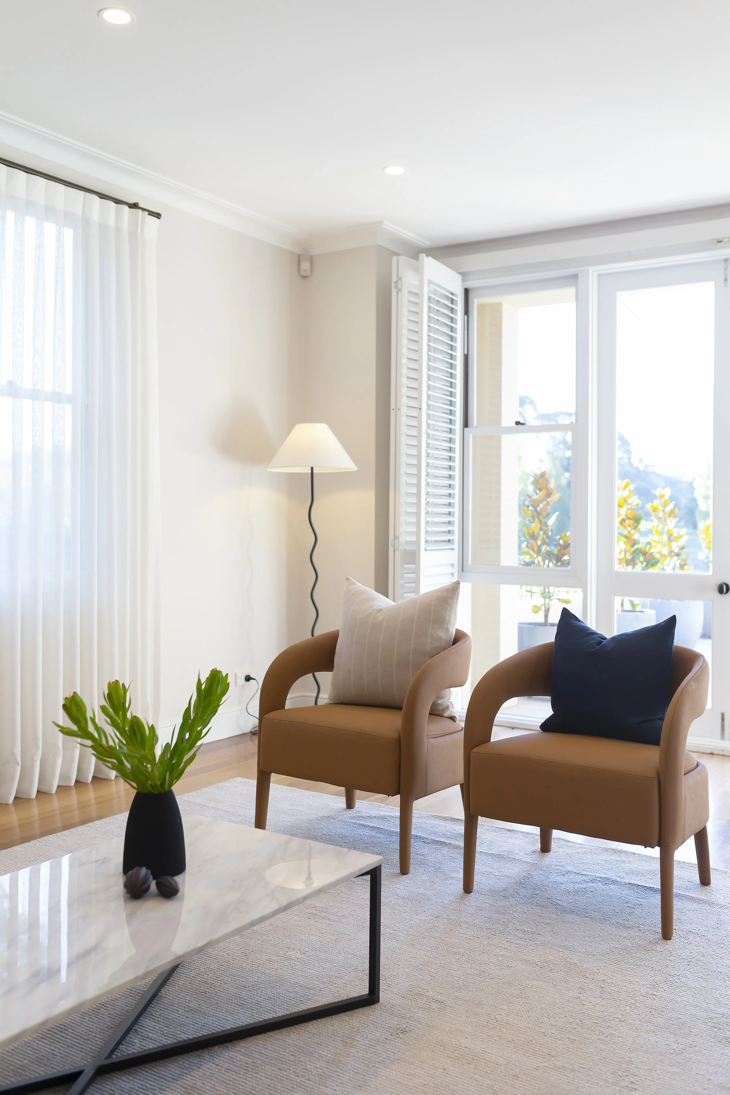 Living room with beige armchairs, a white coffee table with a black vase and green plant, white curtains, and an open window with outside plants.