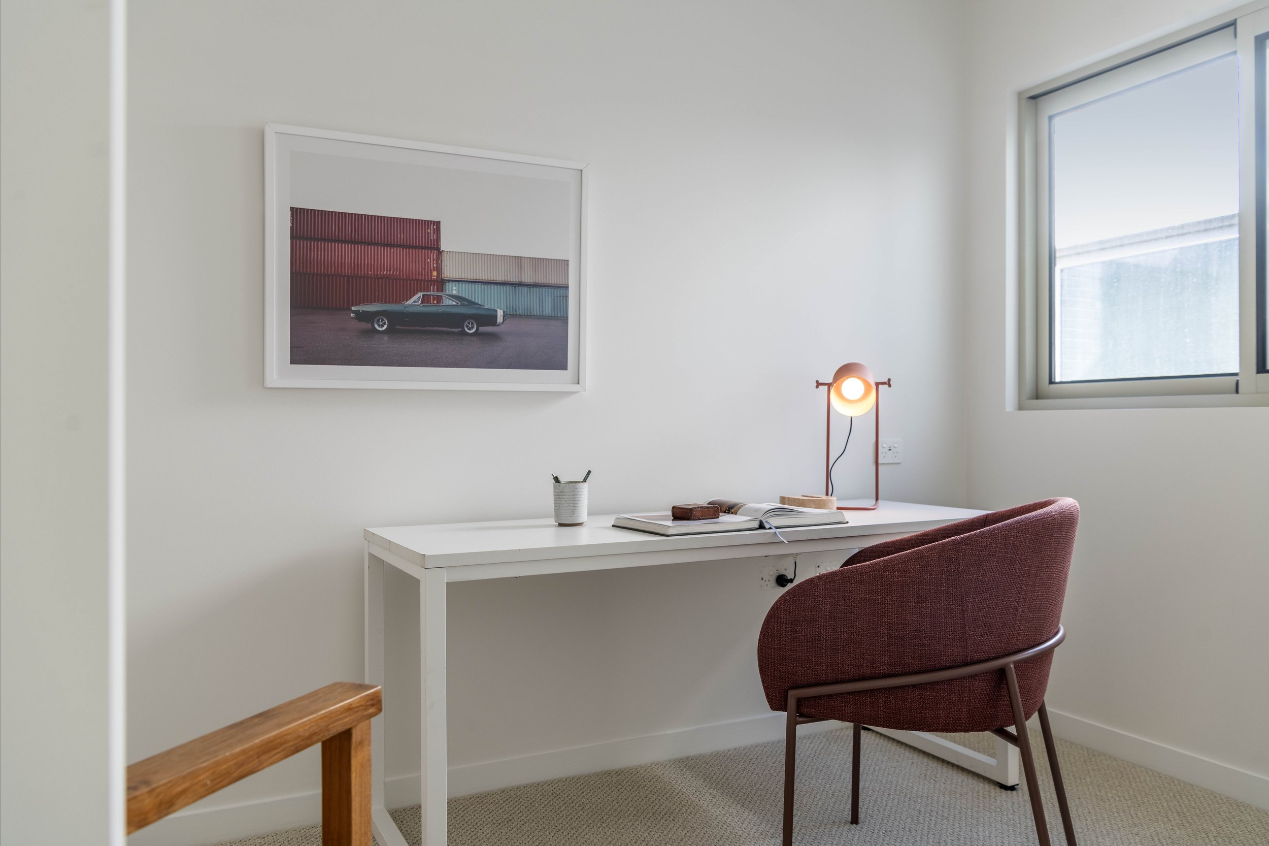 Bright, minimalist home office with white desk, red chair, and a window with a view outside. Wall art features a vintage car against colorful shipping containers. Small lamp, open book, and pen holder on the desk.