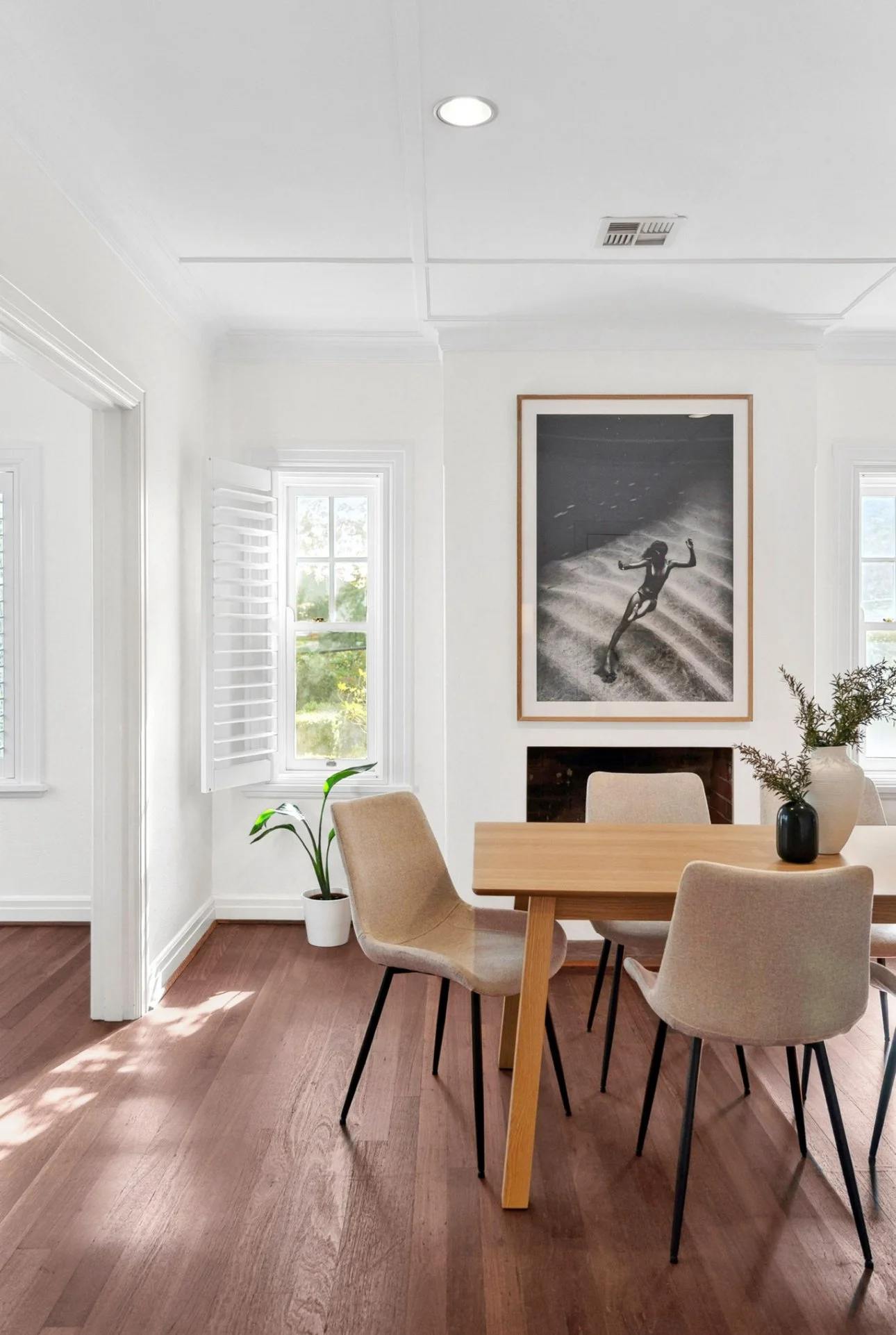 Dining room with wooden table and beige chairs, black and white beach photograph on white wall, potted plant near window, sunlight streaming through window.