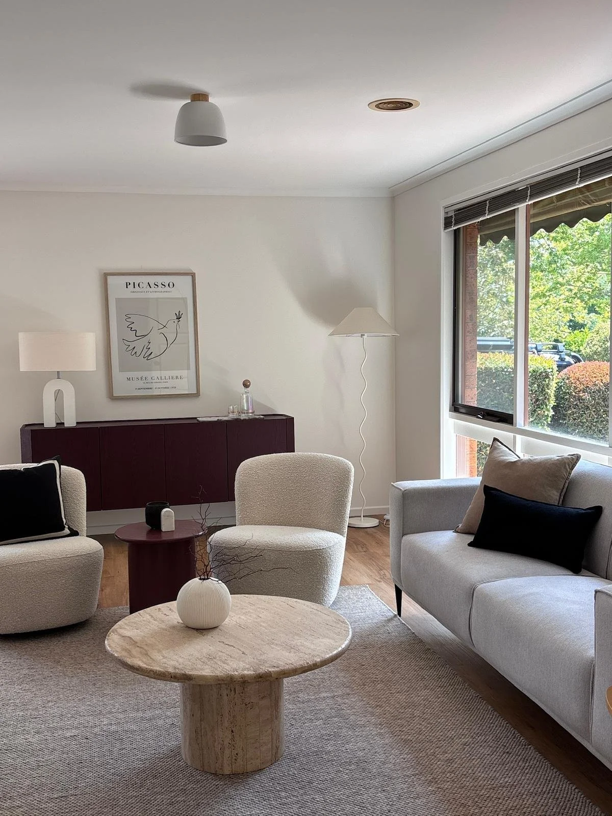 Bright living room with white walls, large window with blinds, white sofa with pillows, beige armchair, round wooden coffee table with a vase, and a dark red sideboard with decorative items and framed artwork on the wall.