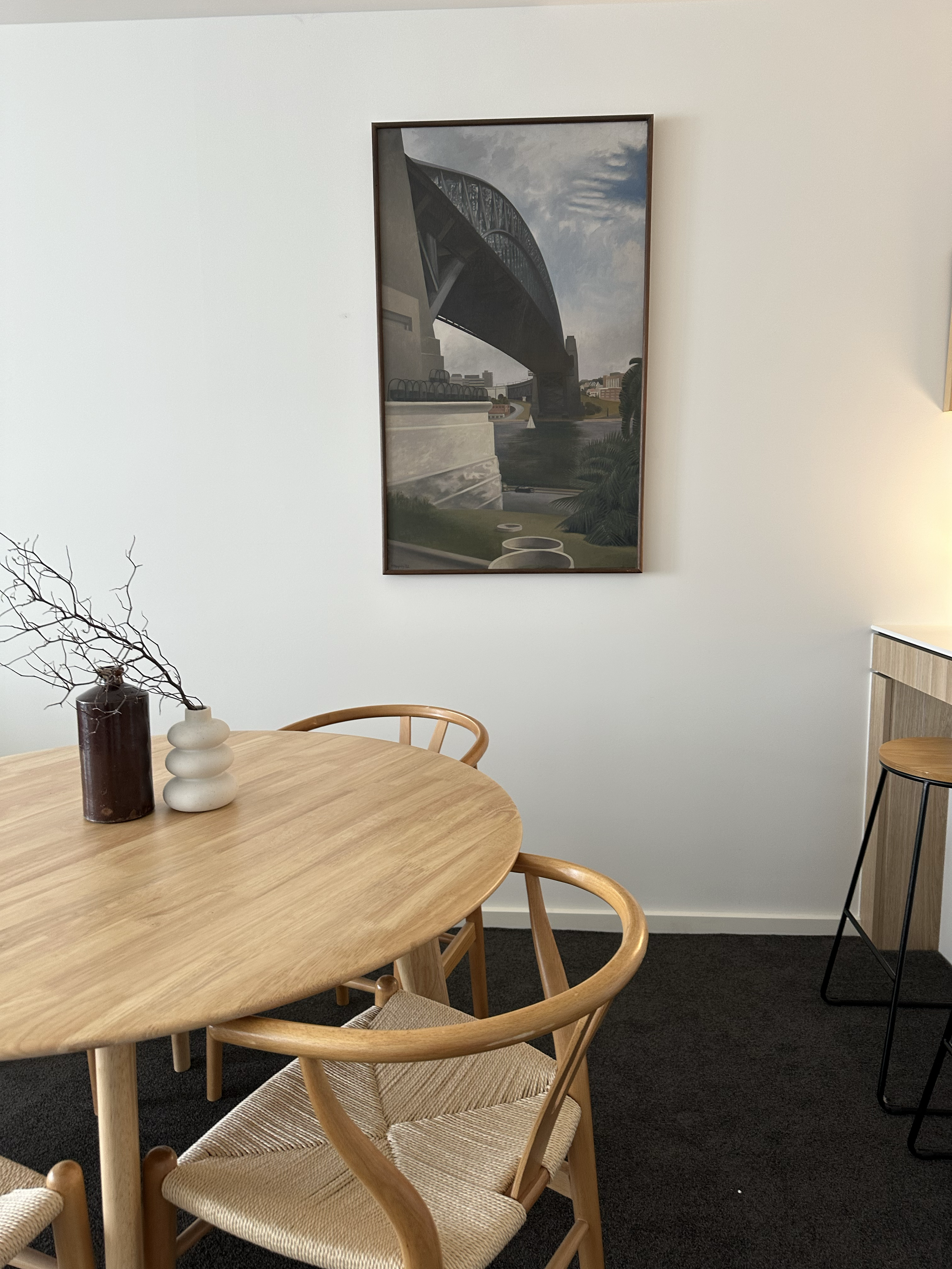 A cozy dining area with a light wooden table and matching chairs, a decorative vase with branches, and framed picture of a bridge on the wall.