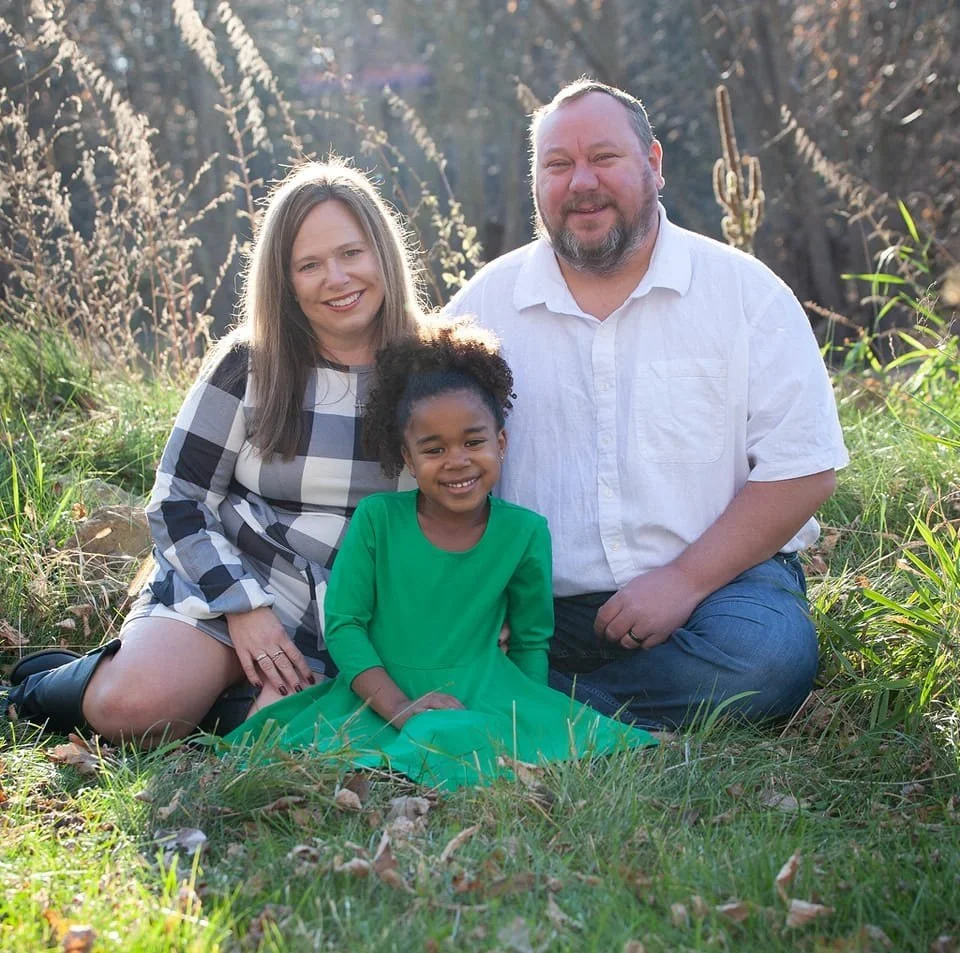 Jeff Fasbender family of three sitting outdoors on grass, smiling at the camera, with a background of trees and sunlight.
