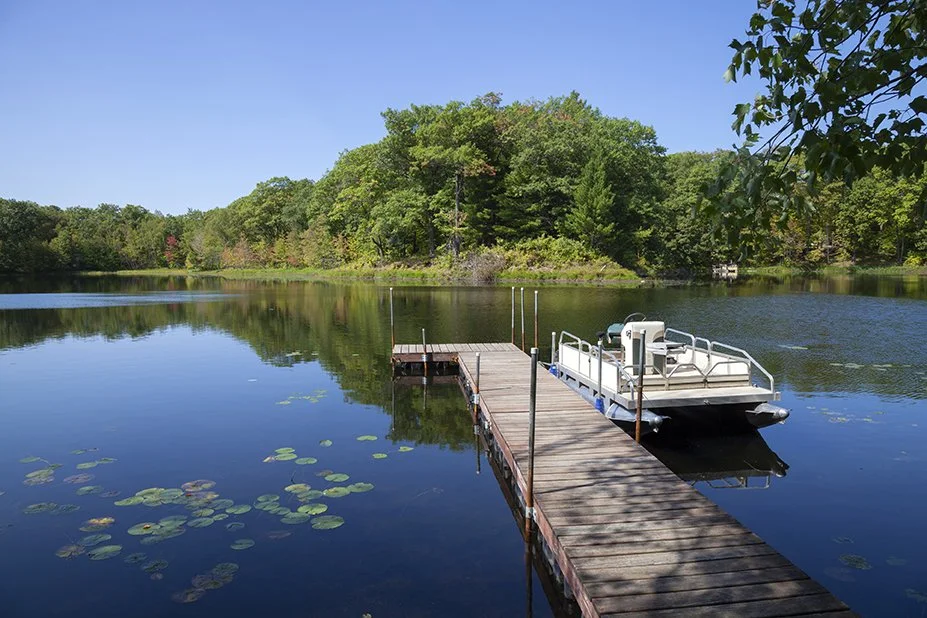 A wooden dock extends into a calm lake with lily pads, a white boat tied to the dock, surrounded by green trees under a clear blue sky.