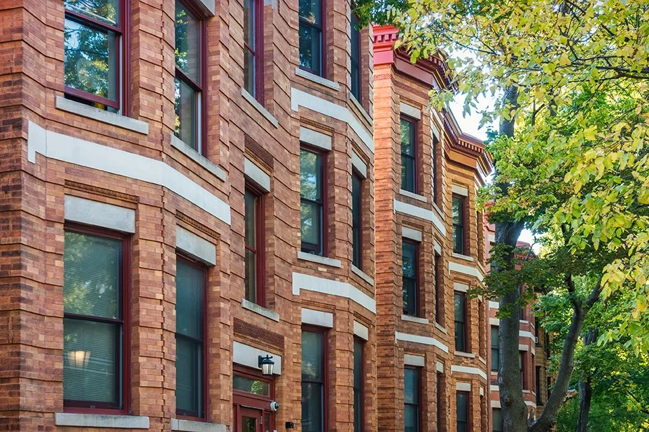 Brick apartment building with windows, partially obscured by green tree leaves.