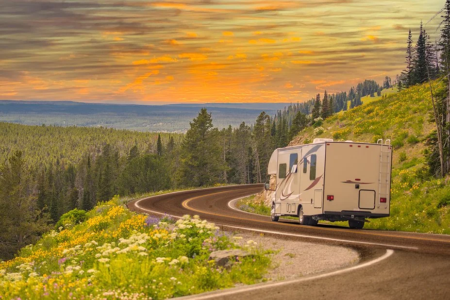 A white RV traveling along a winding mountain road with wildflowers on the roadside and a forested landscape under a colorful sunset sky.