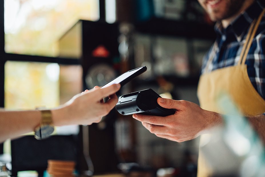 A customer paying with a smartphone at a store counter, with a cashier holding a card reader near the phone.