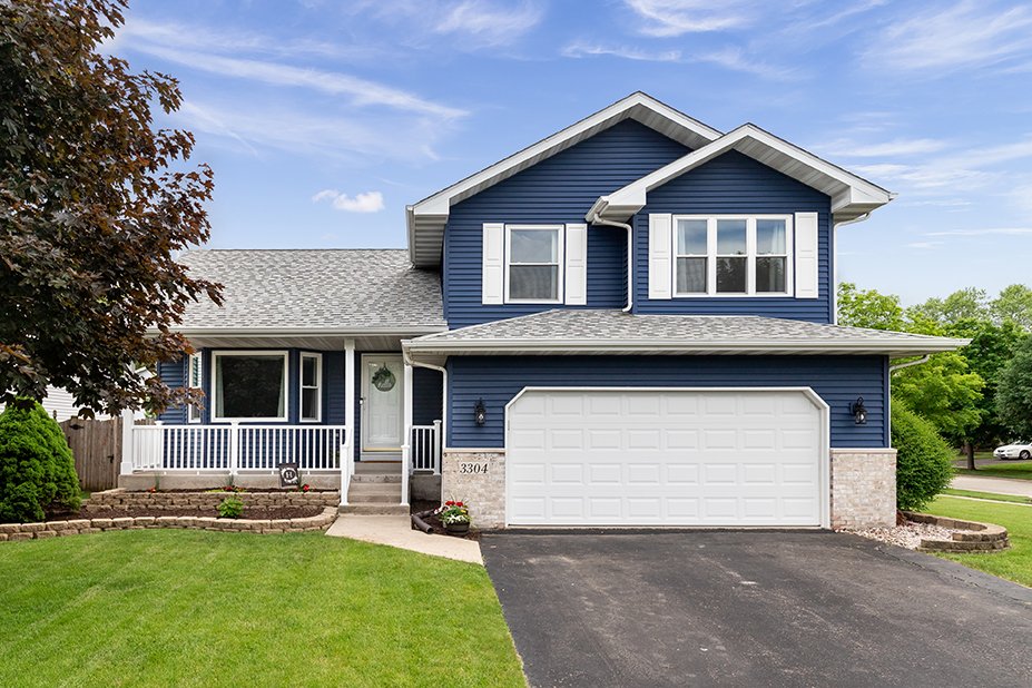 A two-story blue house with white trim, a two-car garage, and a front porch with steps surrounded by a well-maintained lawn.