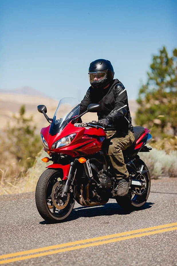 Motorcyclist wearing a helmet and black jacket riding a red sport motorcycle on a road with a clear sky and desert landscape in the background.