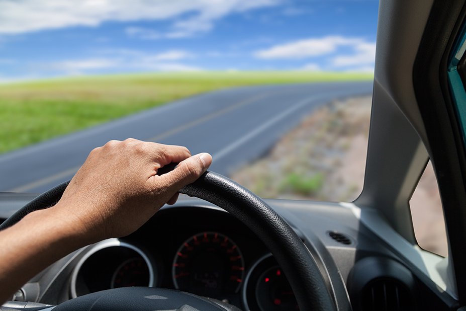 A person's hand on a steering wheel while driving on a rural road with green fields and a blue sky visible outside the car window.