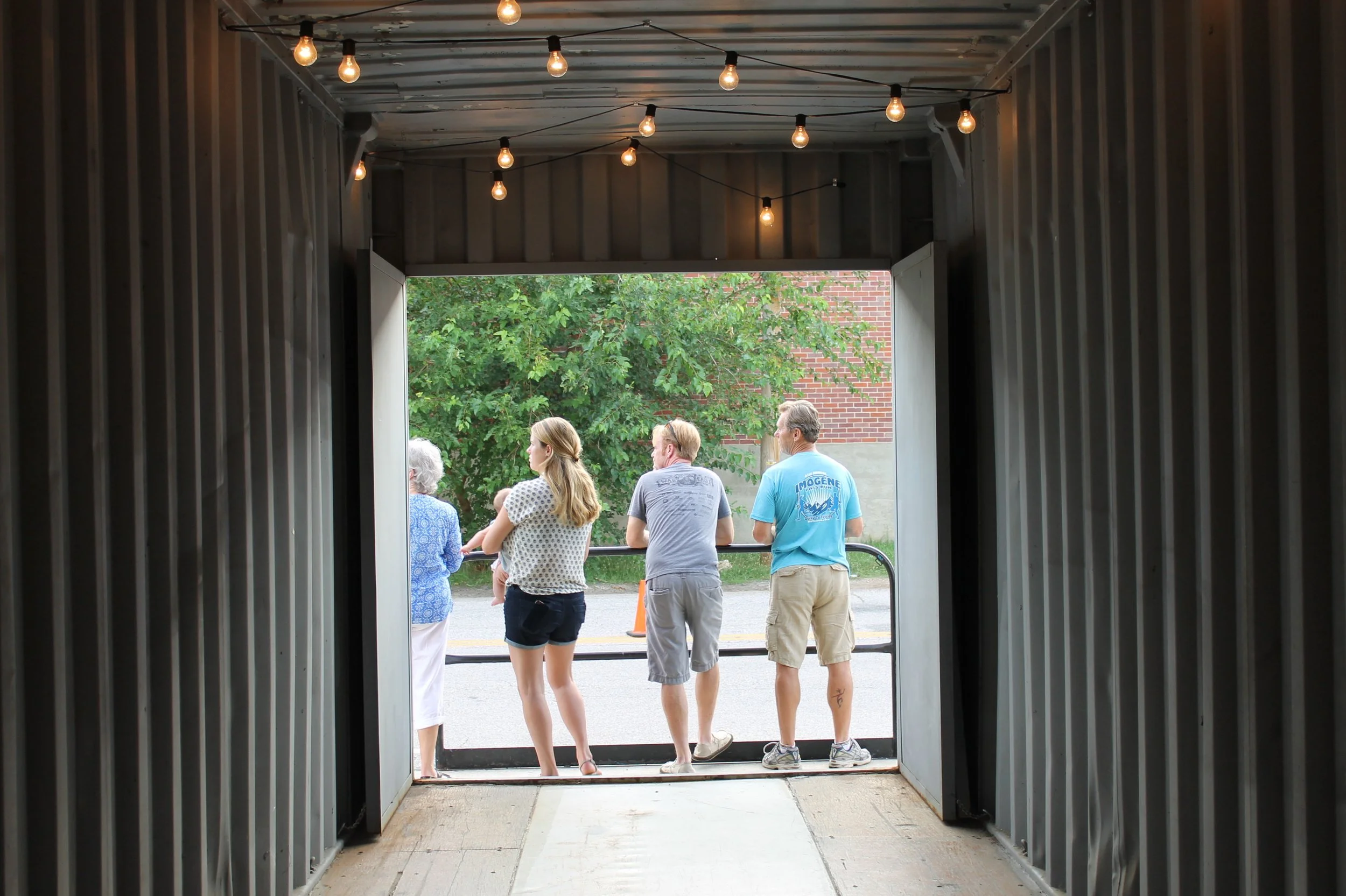 Slowpoke Studio’s website redesign for Wiseacre Brewing Company. People standing behind a barricade, seen from inside a dark tunnel with string lights overhead, looking out onto a tree and brick building.