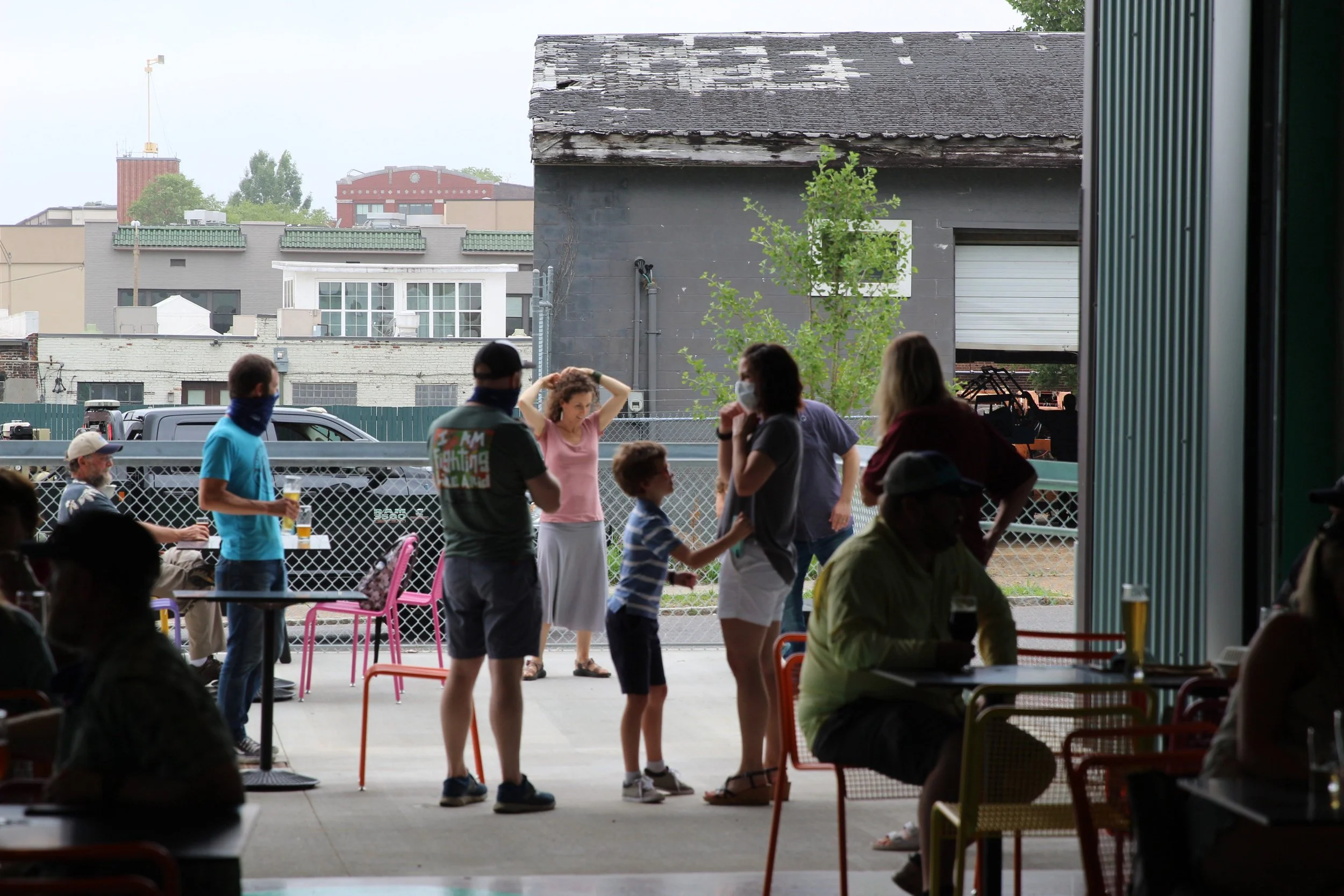 Slowpoke Studio’s website redesign for Wiseacre Brewing Company. People socializing some standing and some sitting, with a building, parked cars, and a chain-link fence in the background.