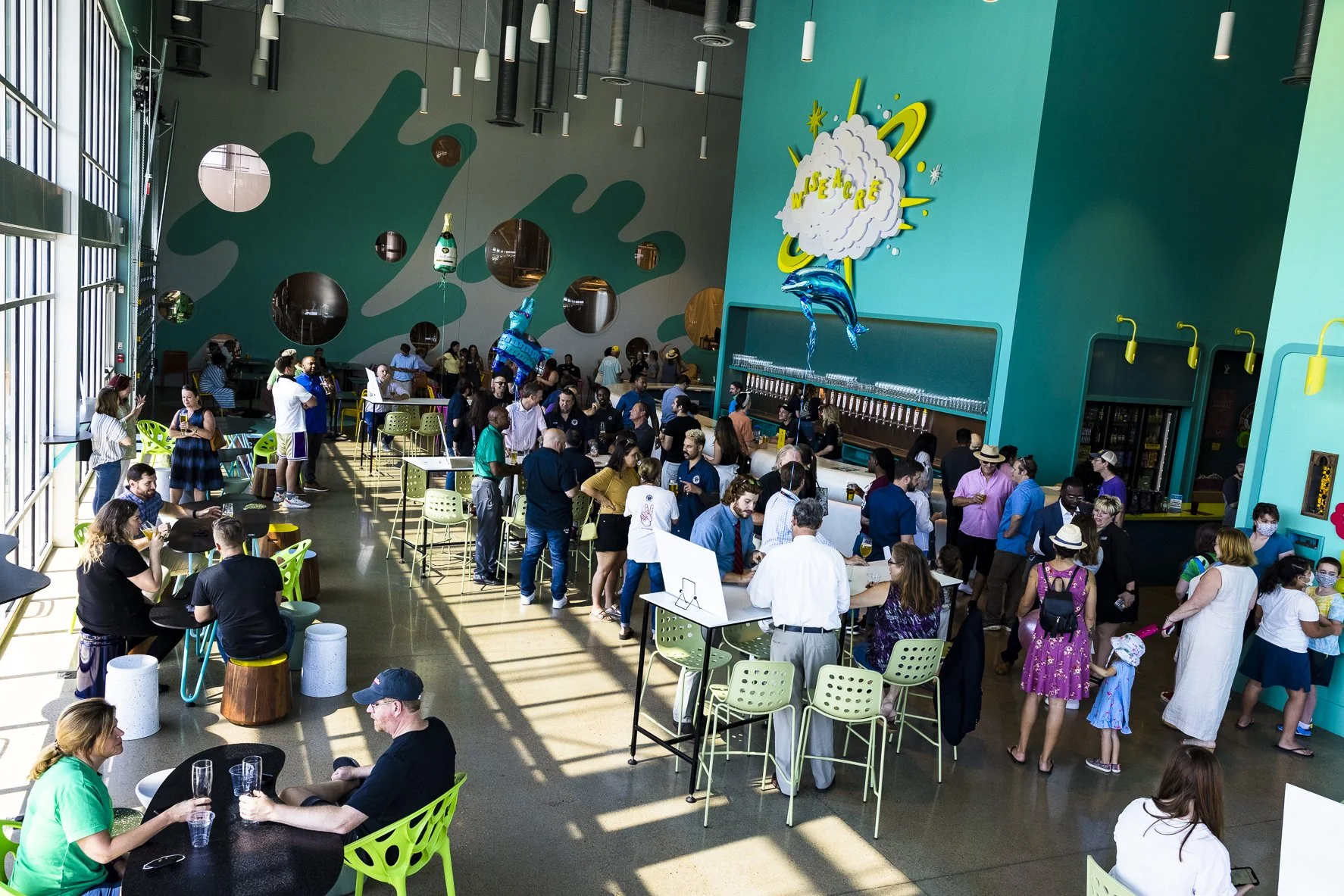 Slowpoke Studio’s website redesign for Wiseacre Brewing Company. People gather inside a colorful, modern brewery or bar with teal walls, decorated with playful cloud motif, and large windows letting in natural light.