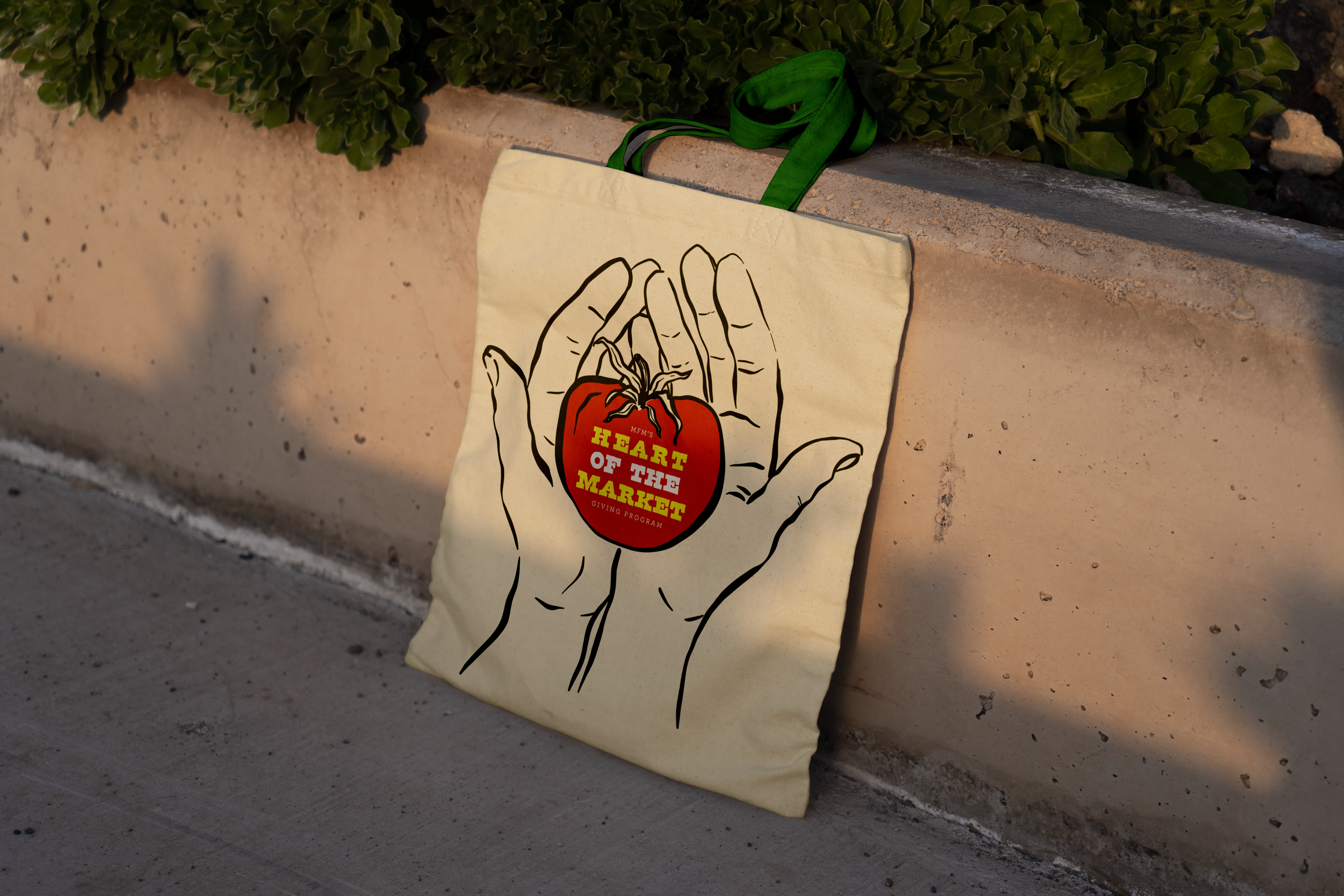 Slowpoke Studio refreshed the Memphis Farmers Market merch design adding anew revenue stream. A beige tote bag with illustration of hands holding a tomato  that reads 'Heart of the Market' in bold yellow text.