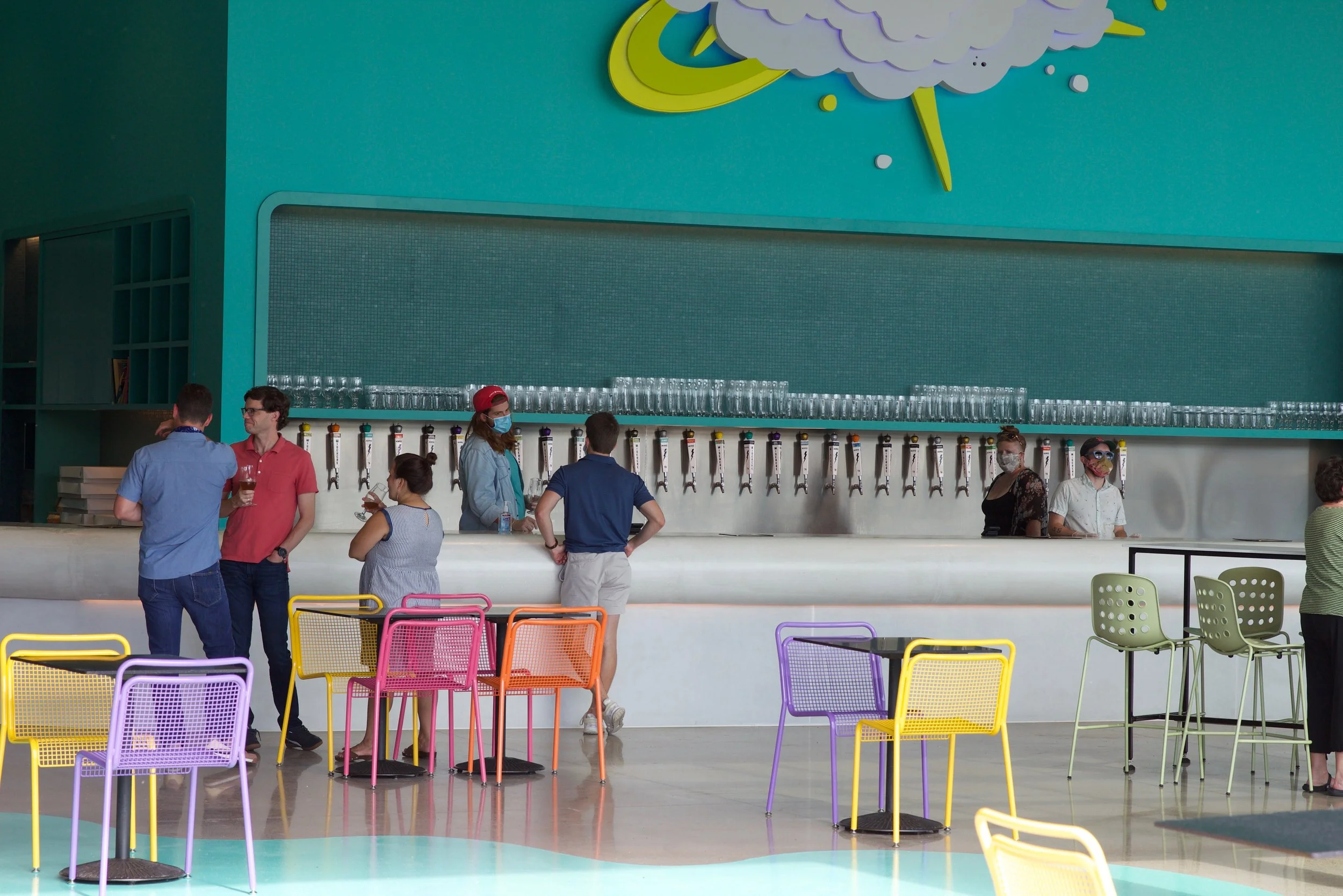 Slowpoke Studio’s website redesign for Wiseacre Brewing Company. People at a bar counter with colorful chairs in a modern space. There are multiple beer taps and glasses above the counter, and some people are wearing masks.