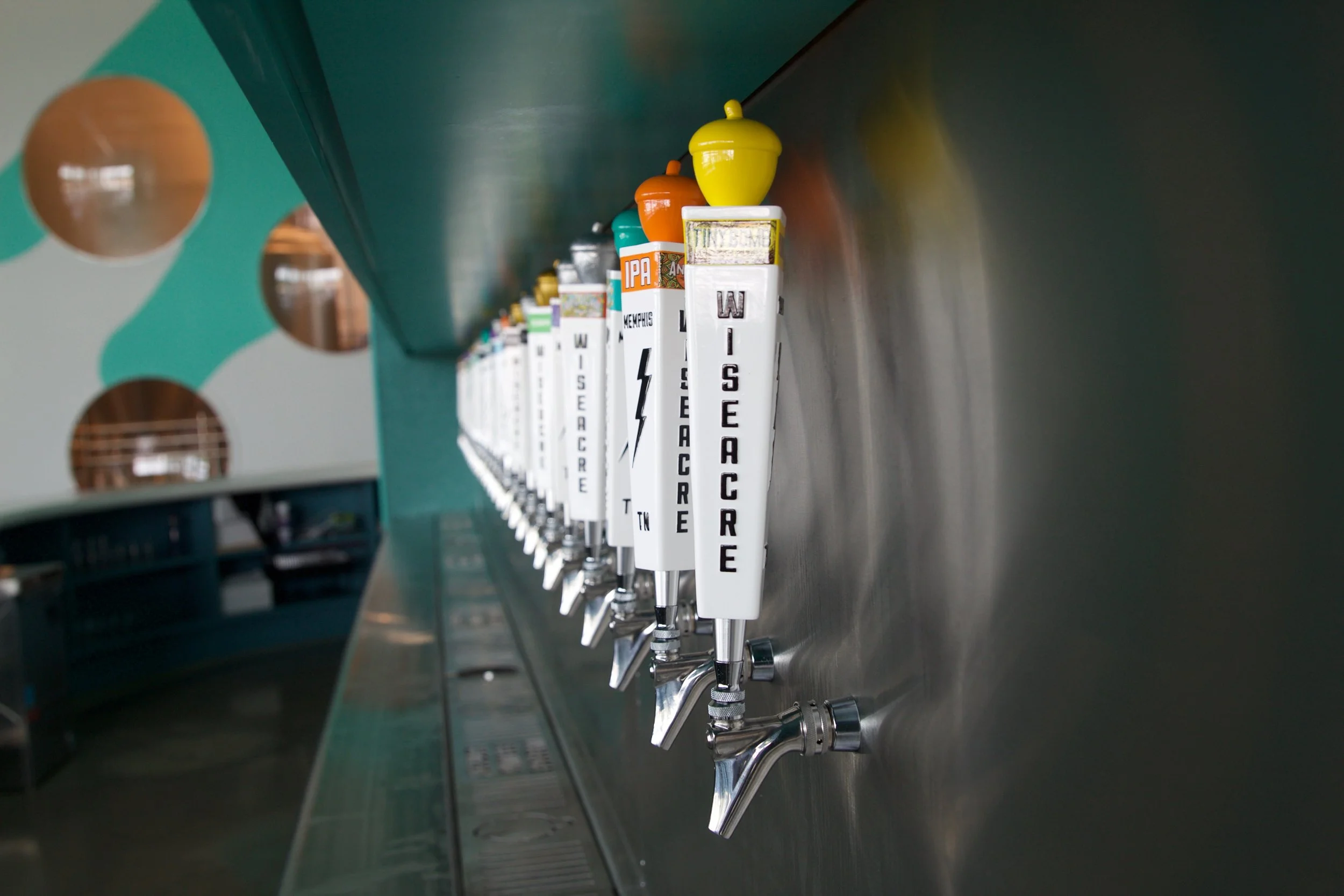 Slowpoke Studio’s website redesign for Wiseacre Brewing Company. Close-up of beer taps with colorful handles labeled with different beer types, mounted on a stainless steel bar in a brewery.