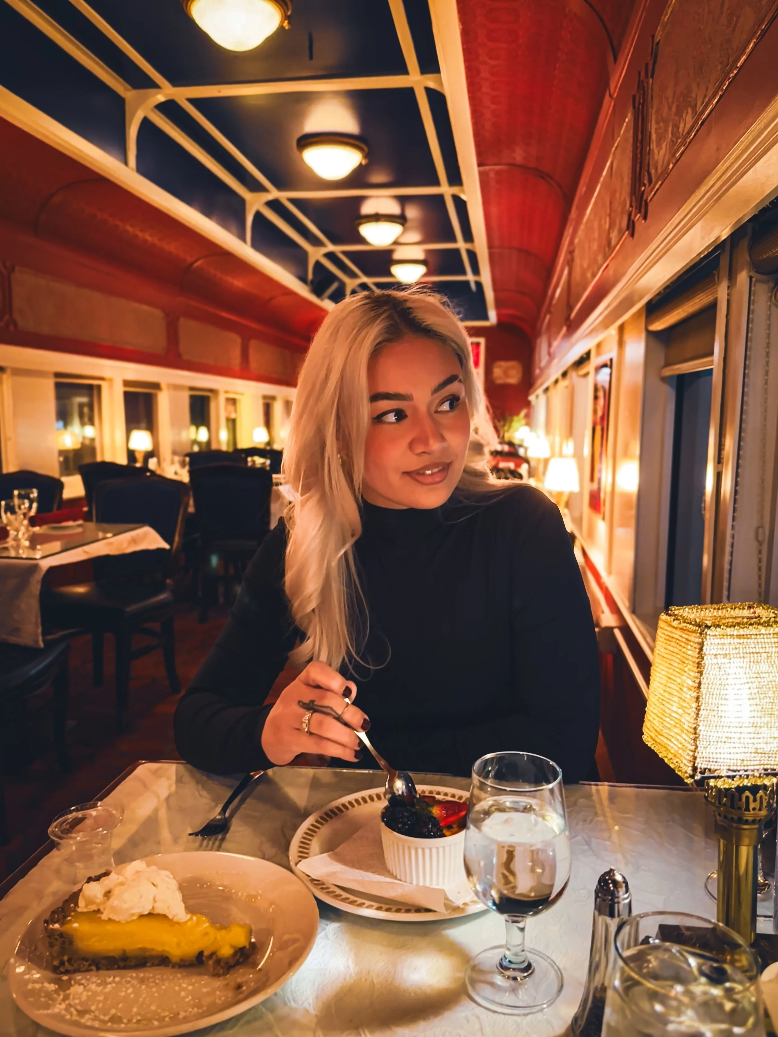 A woman with blonde hair and a black top sitting at a restaurant table with desserts and a glass of water, in a warmly lit dining room.