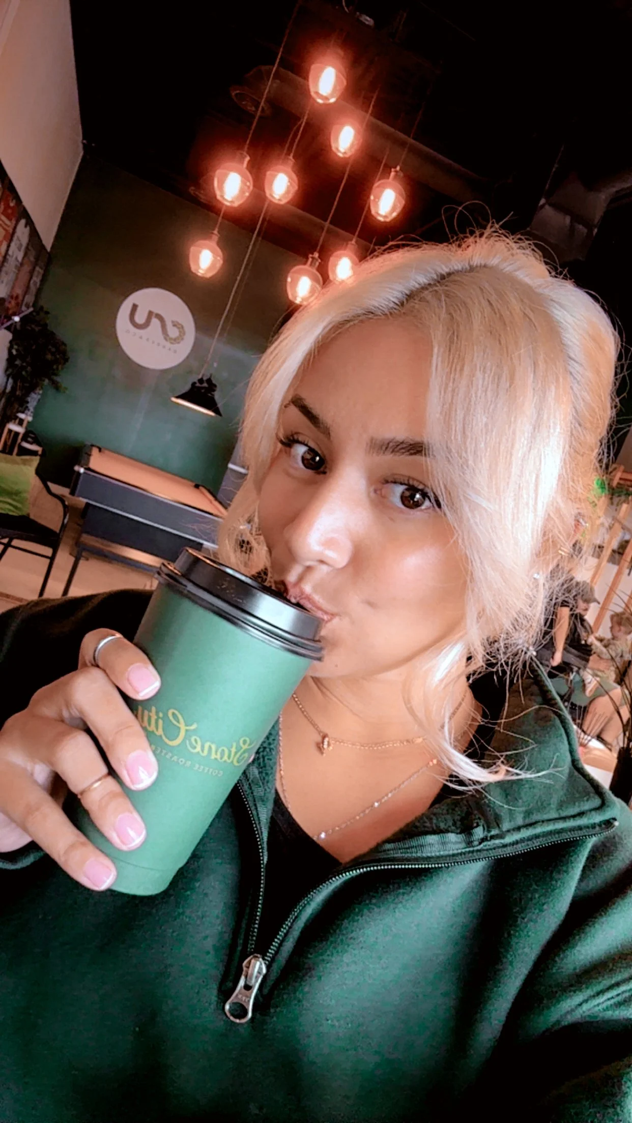 A woman with blonde hair drinking from a green travel mug in a coffee shop with hanging lights and a pool table in the background.