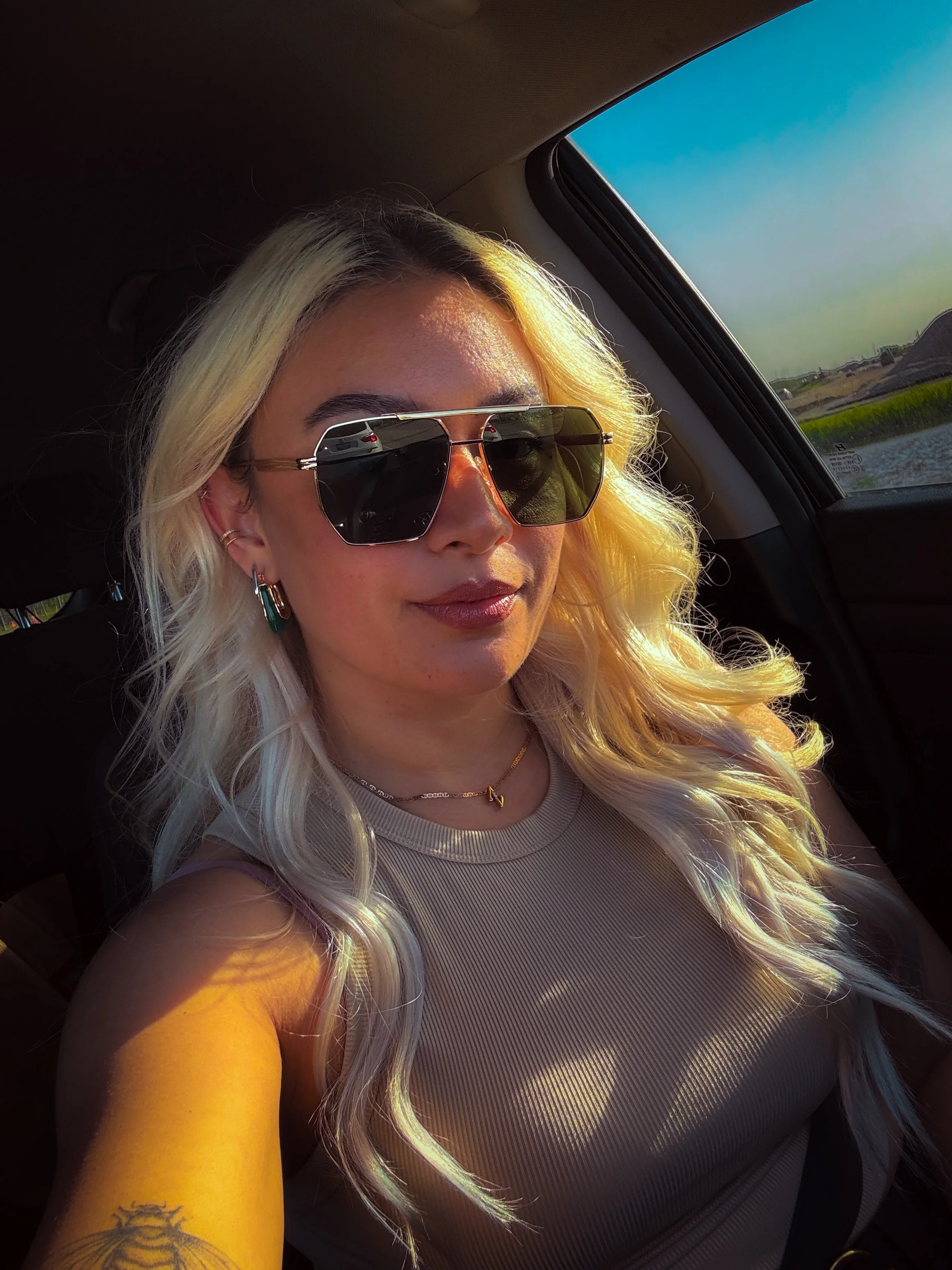 A woman with long blonde wavy hair wearing large sunglasses and jewelry taking a selfie inside a car with sunlight on her face.