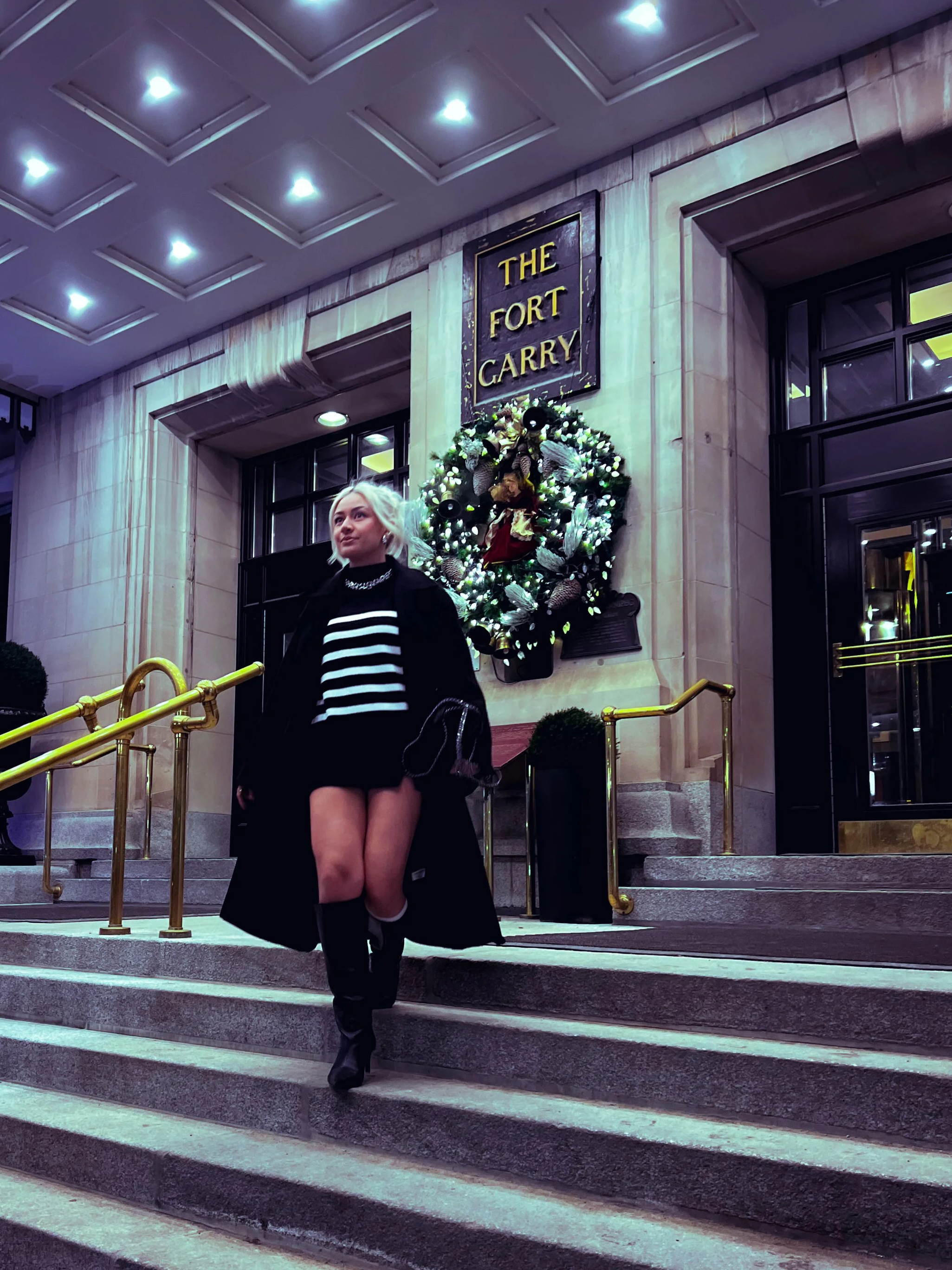 A blonde woman with a black coat, striped shirt, and tall black boots descending stairs outside The Fort Garry hotel, decorated with a Christmas wreath and sign.