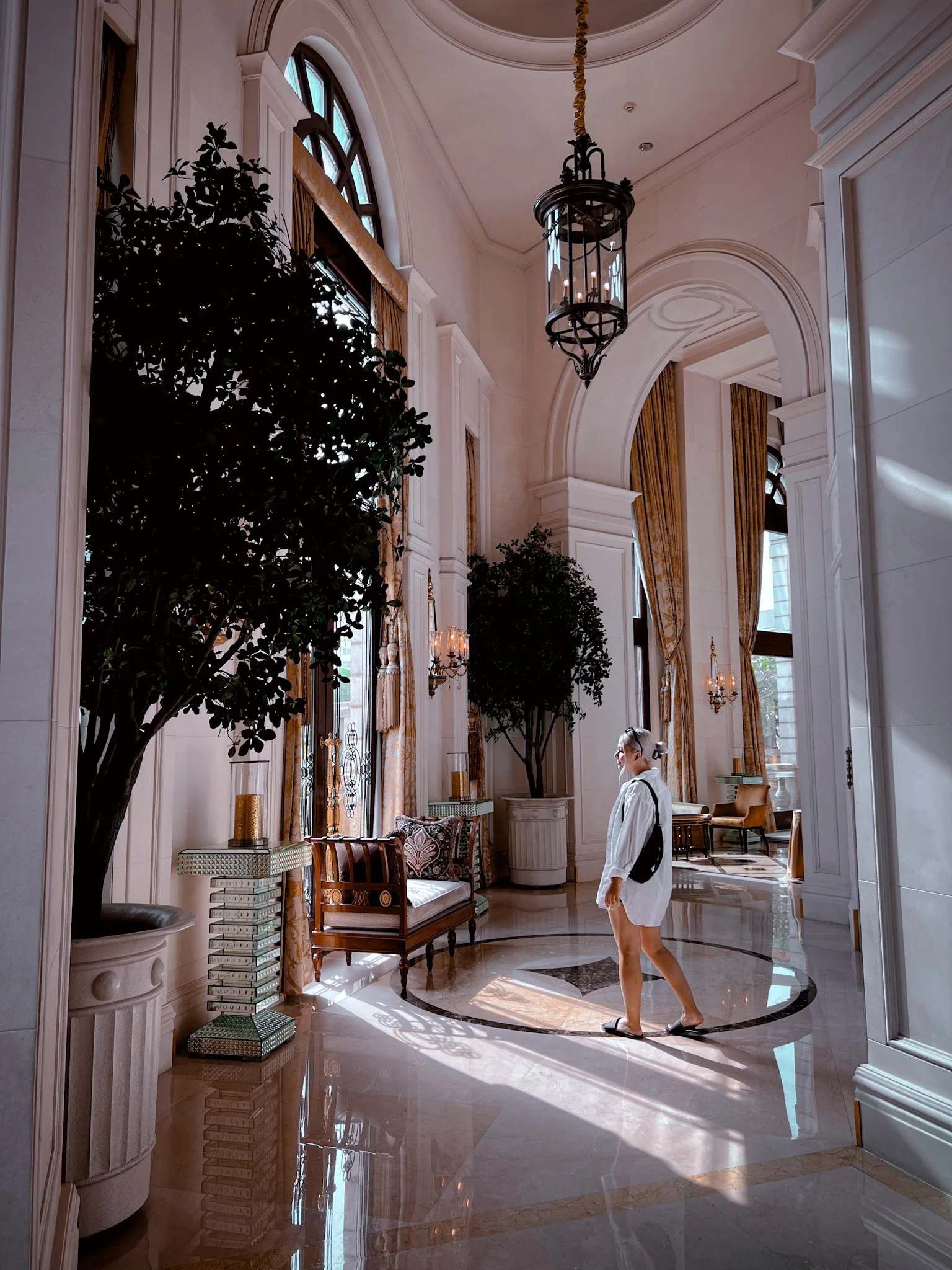 A woman walking in a luxurious, spacious hotel lobby with high ceilings, large windows with golden curtains, and elegant decor.