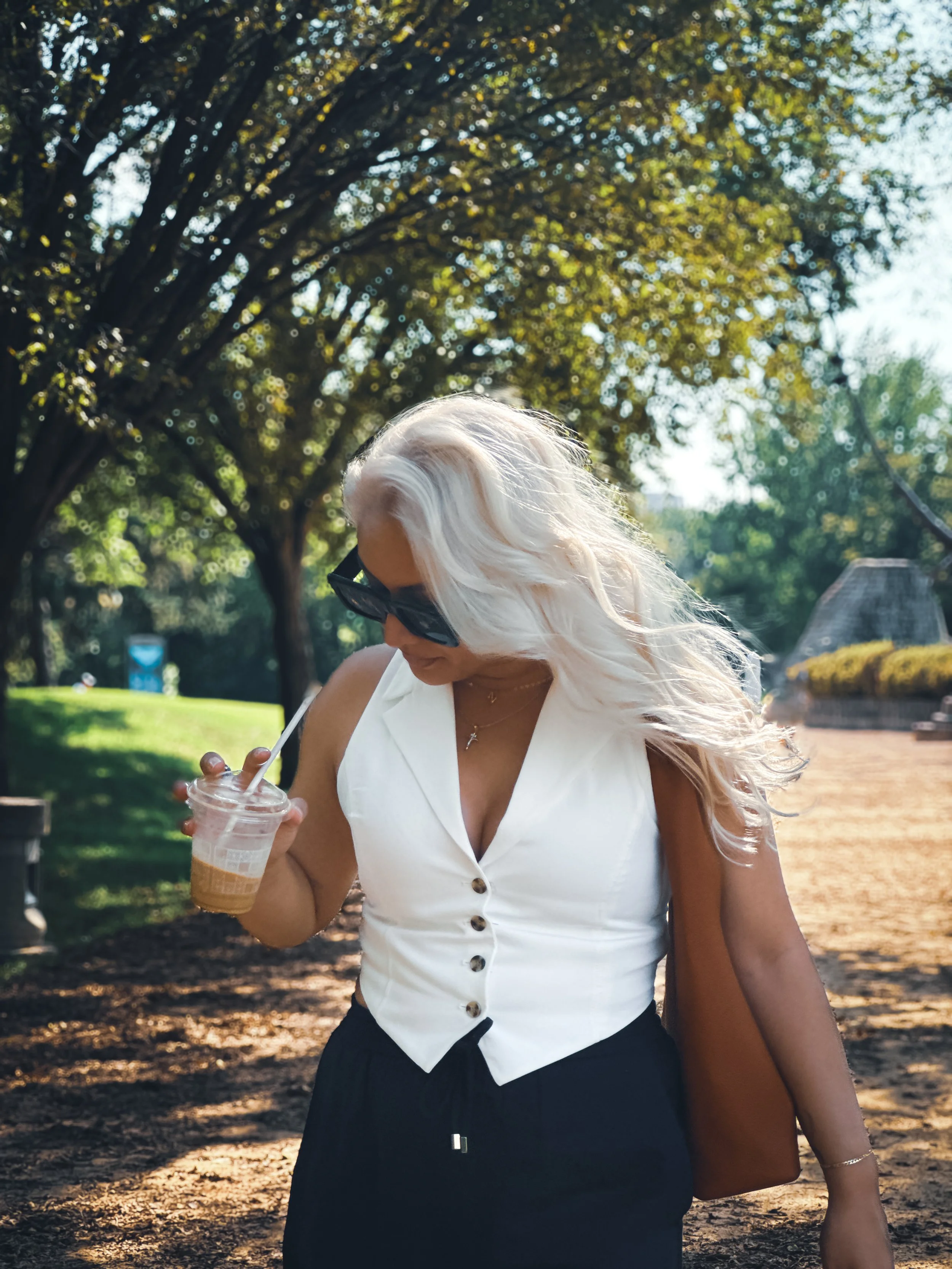 A woman with long, wavy blonde hair, wearing sunglasses, a sleeveless white button-up top, and black pants, holding an iced coffee in a clear cup while walking outdoors on a sunny day with trees and a park in the background.
