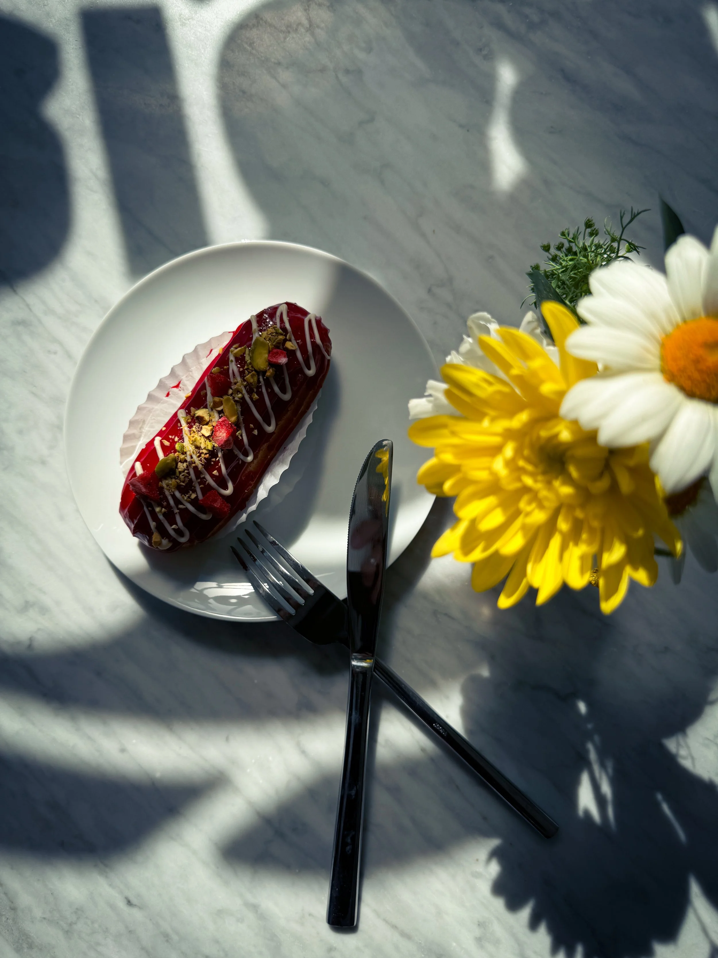 A white plate with a decorated pastry, topped with white drizzle and crushed nuts or dried fruit, on a marble table. There are a fork and a knife crossed on the plate, and a small bouquet of white and yellow flowers nearby.