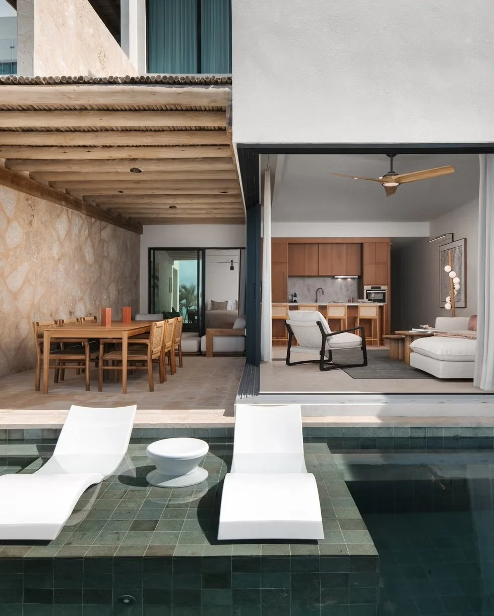 A modern house with a pool area, featuring a wooden dining table with six chairs, a living room with white couches, a ceiling fan, a kitchen with wooden cabinets, and poolside lounge chairs.