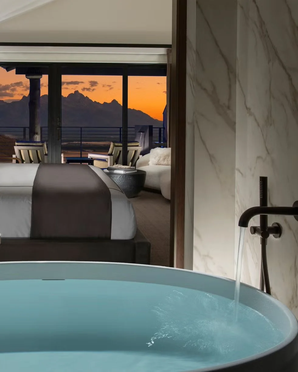 A hotel room with a view of mountains at sunset, featuring a bathtub with water in the foreground and a bedroom with a bed and seating area in the background.