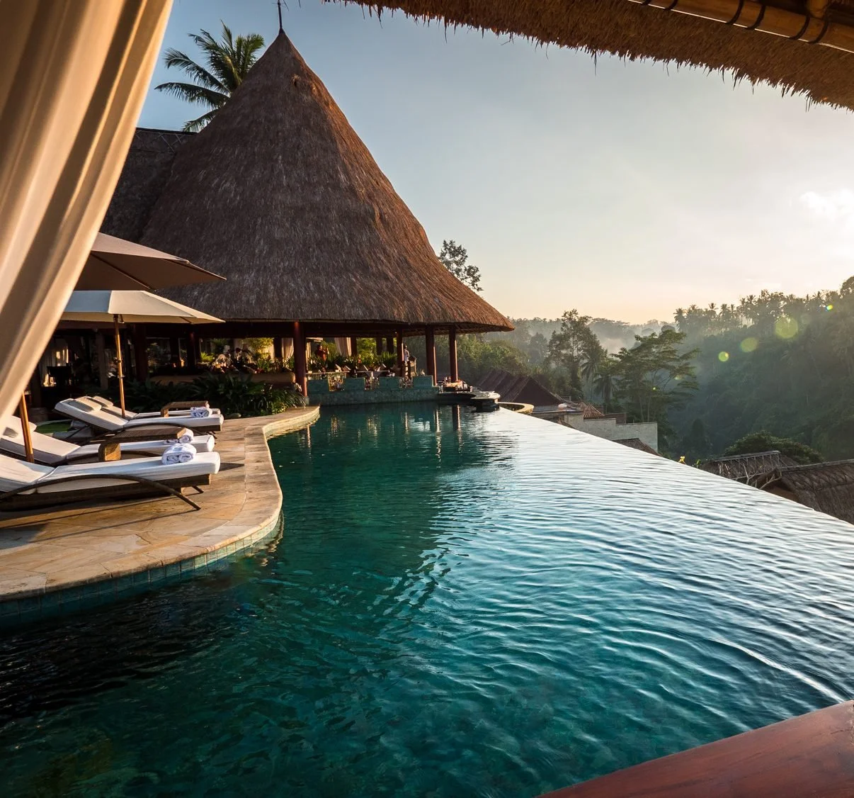 Outdoor patio with a swimming pool, beige sofa with pillows, lounge chairs, and a dining area under a covered roof, surrounded by trees and scenic landscape in the background.