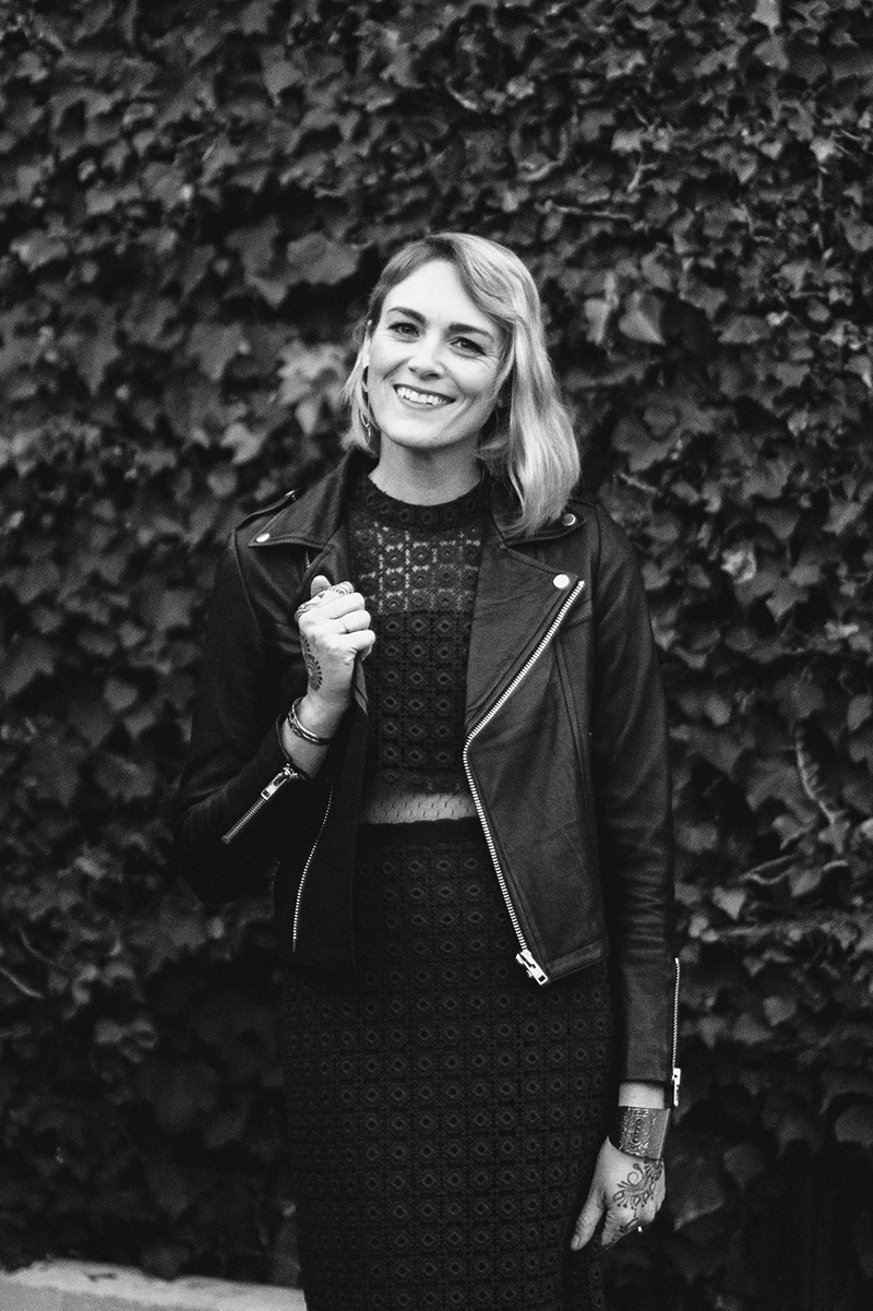 A smiling woman standing outdoors in front of a leafy background, wearing a leather jacket and a lace dress.