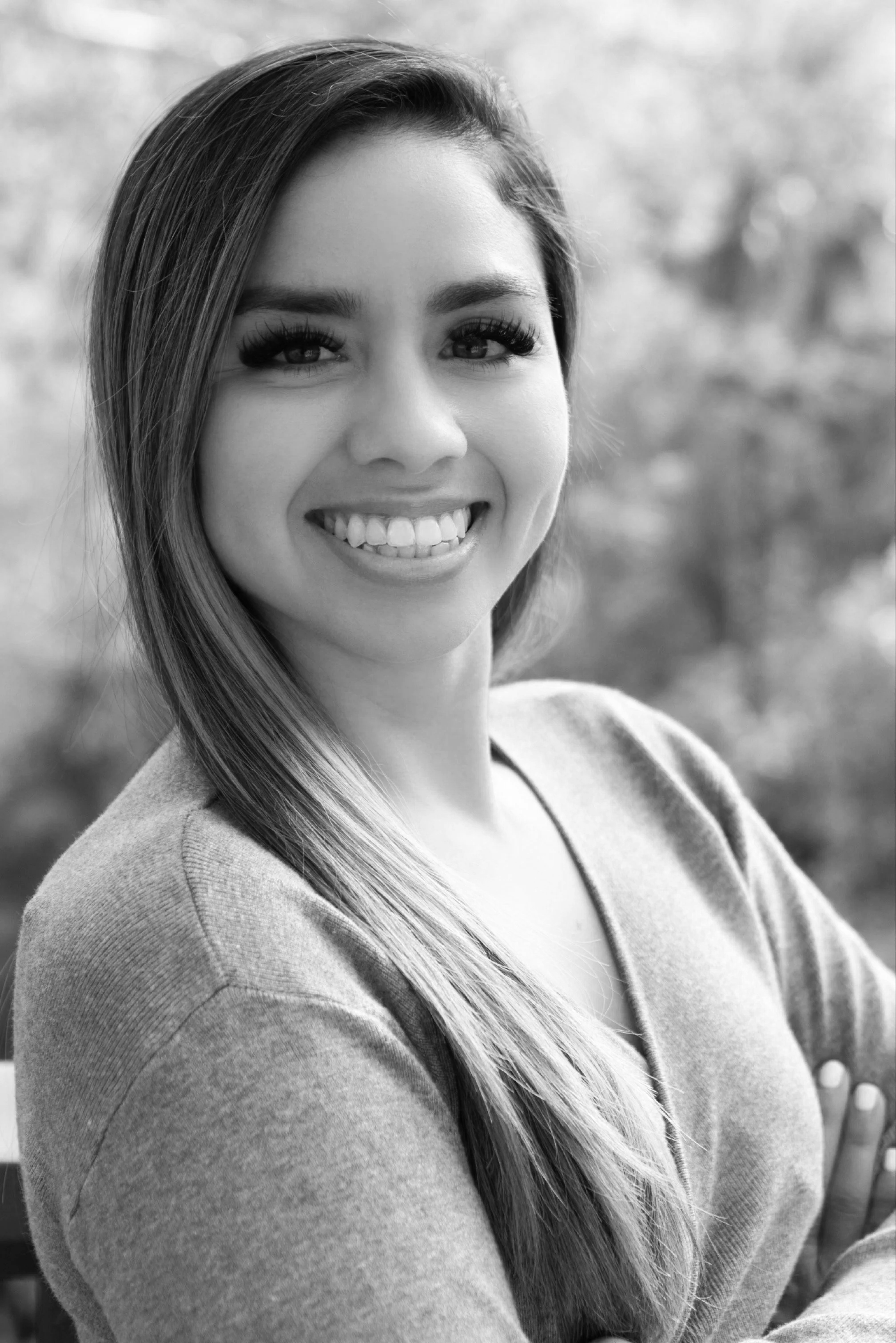 A smiling woman with long hair, wearing a sweater, outdoors with blurred trees in the background.