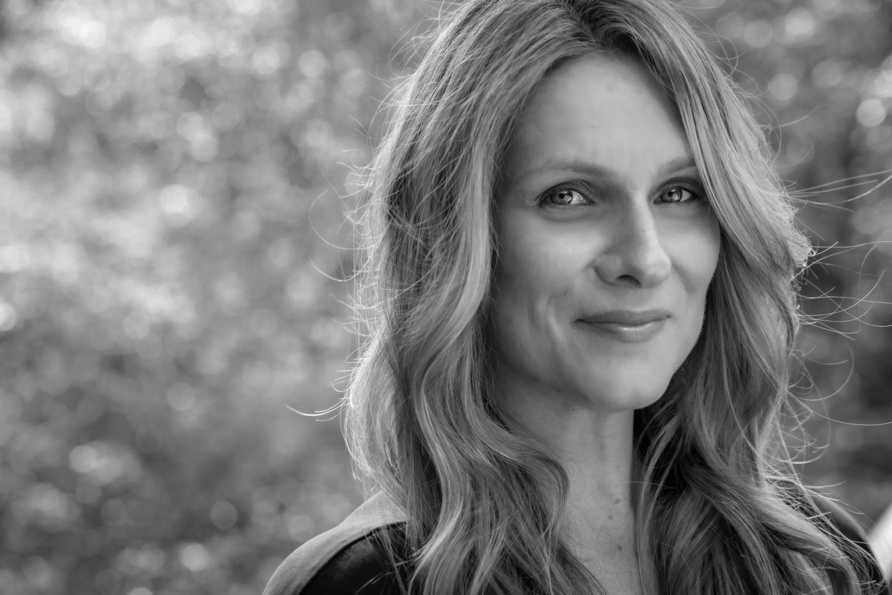 Black and white portrait of a woman with wavy hair smiling slightly, outdoors with blurred background of trees.