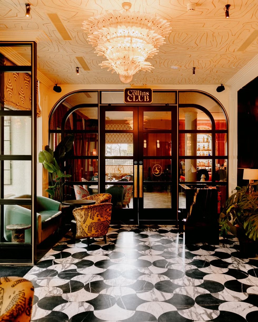 Elegant lobby entrance with a large chandelier, patterned black and white marble floor, glass door marked 'The Collins Club', and plush seating with colorful upholstery.