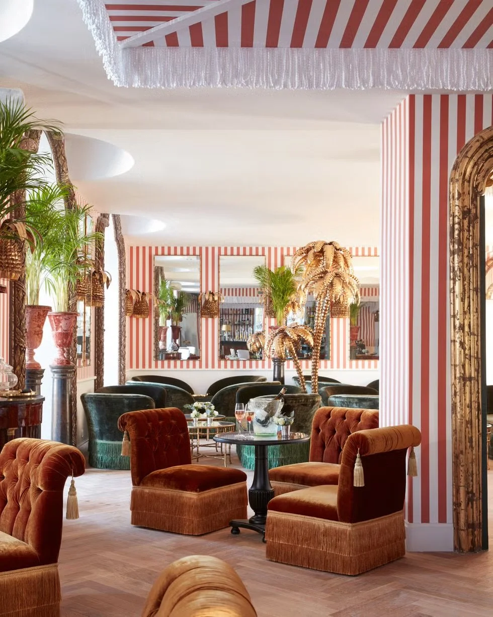A vintage-style lounge with orange velvet chairs, palm trees in decorative planters, striped wallpaper, and a ceiling draped with white sheer fabric.