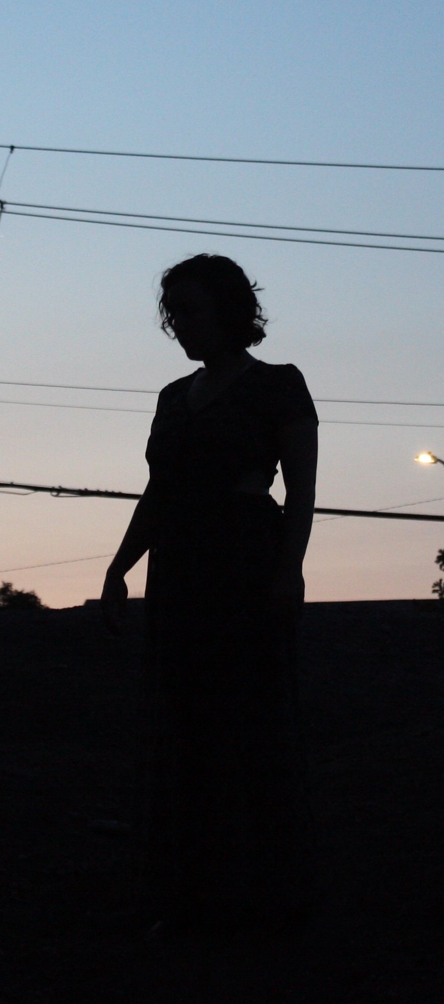 Silhouette of a woman standing outdoors during dusk, with power lines and a streetlight in the background.