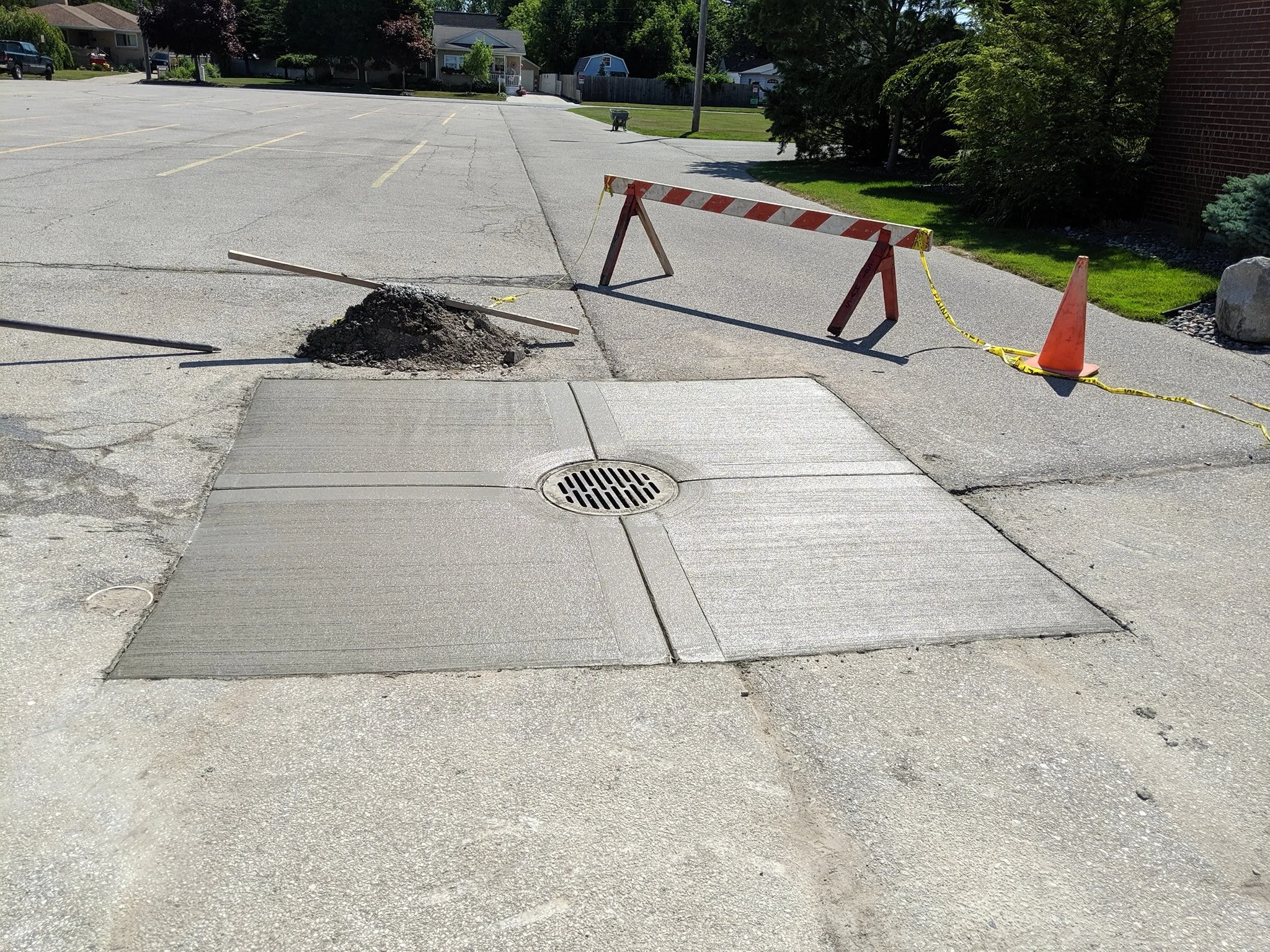 Construction site with a storm drain being repaired, surrounded by cones, a barricade, and caution tape, in a parking lot.