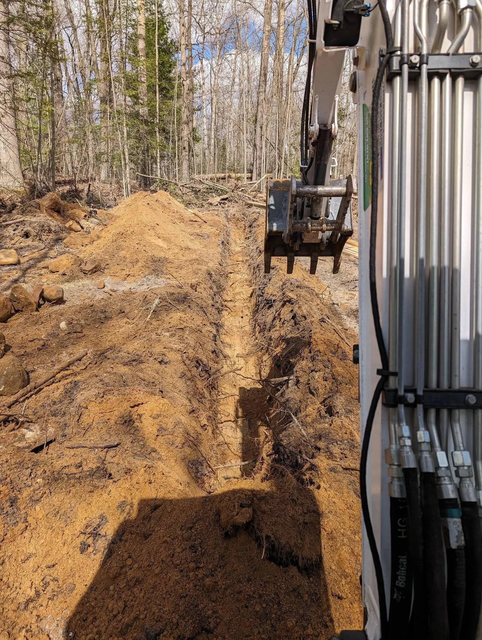 Close-up of a construction site in a forested area during daytime showing an excavator digging a narrow trench in the ground.