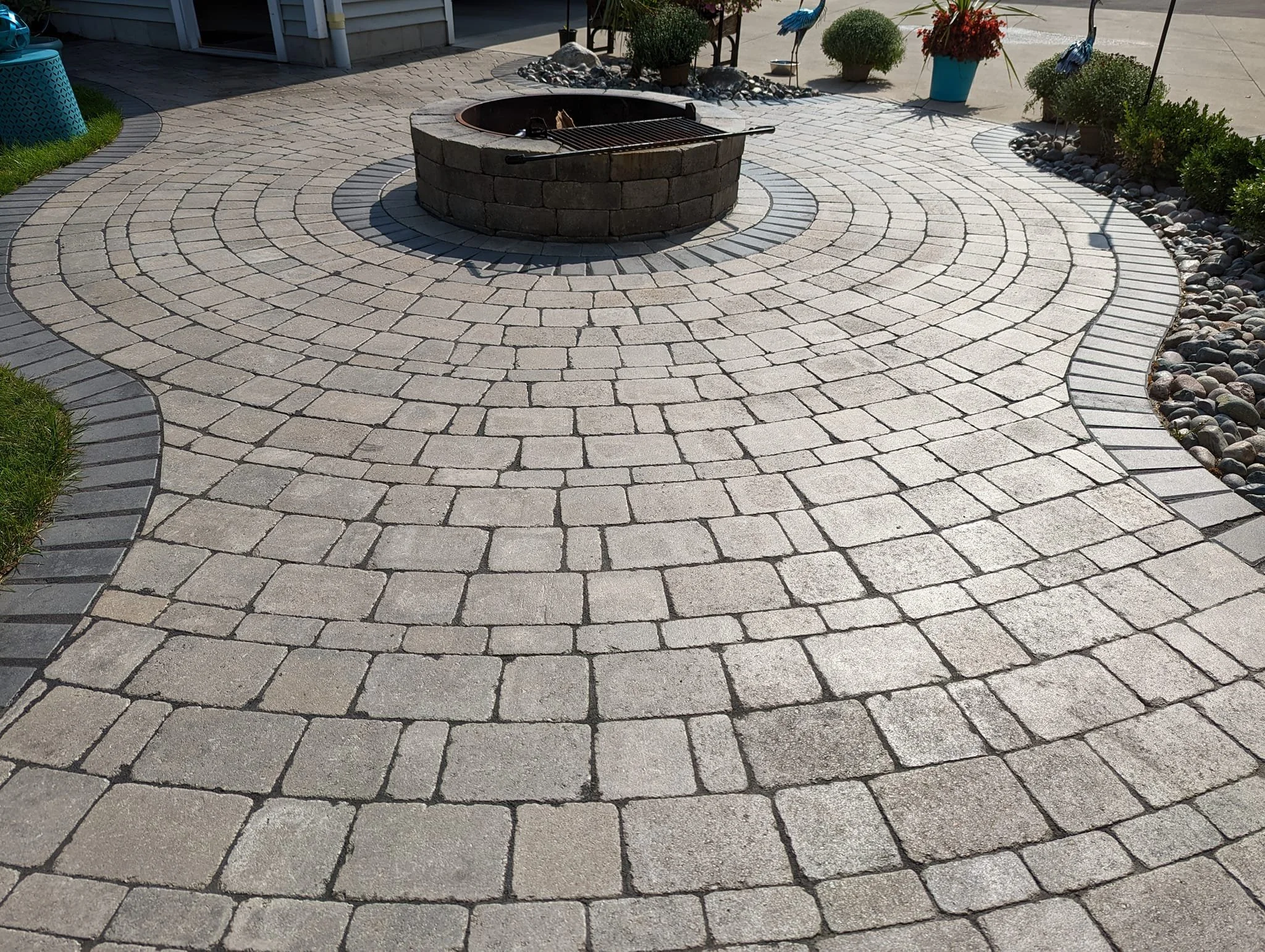 A circular outdoor patio with a fire pit in the center, paved with interlocking stone bricks in concentric circles, and surrounded by landscaped plants and decorative rocks.