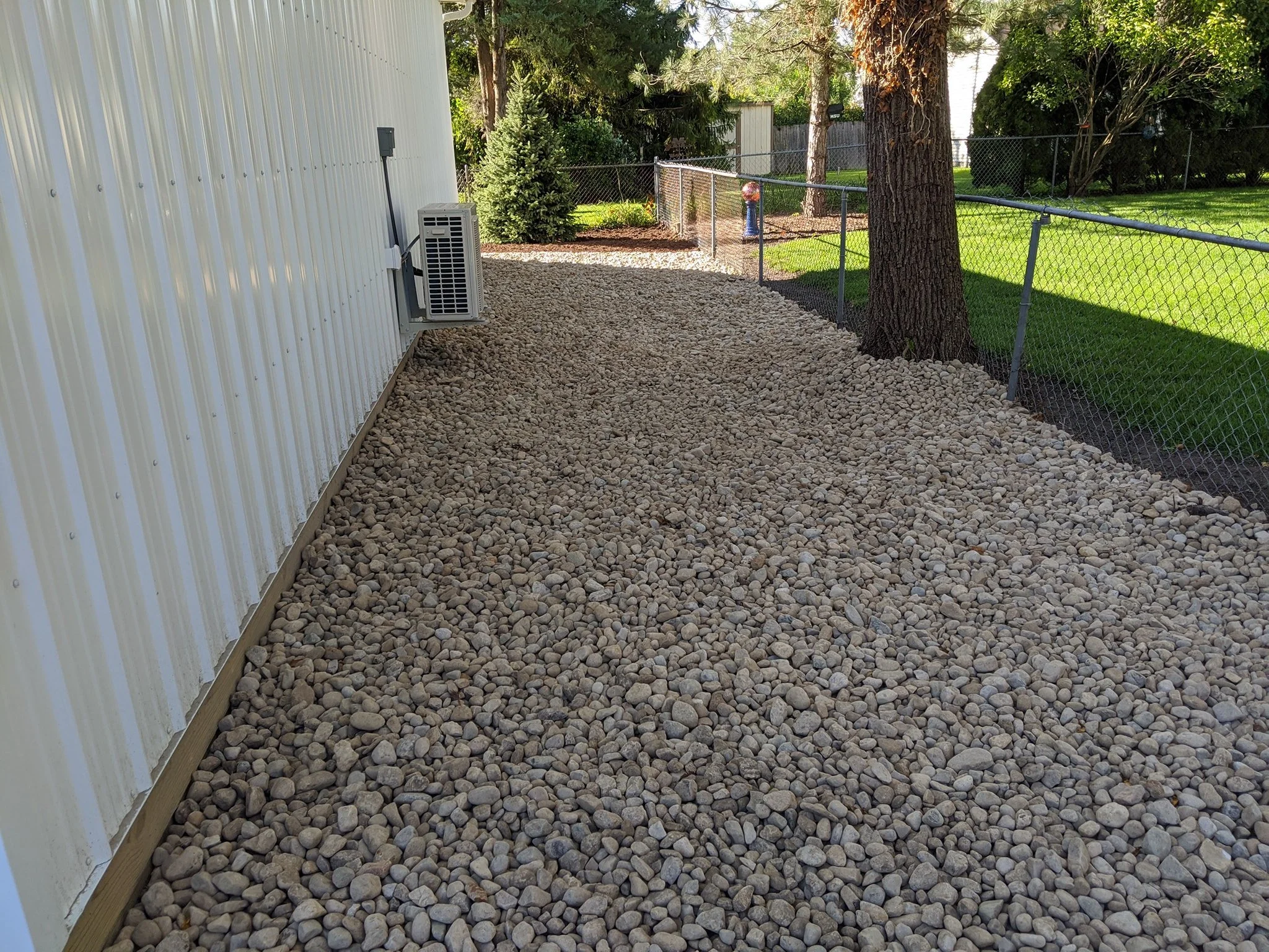 Side yard with ground covered in small gray and beige rocks, a white metal building on the left, a tree on the right, and a chain-link fence dividing the yard with some green grass, shrubs, and trees in the background.