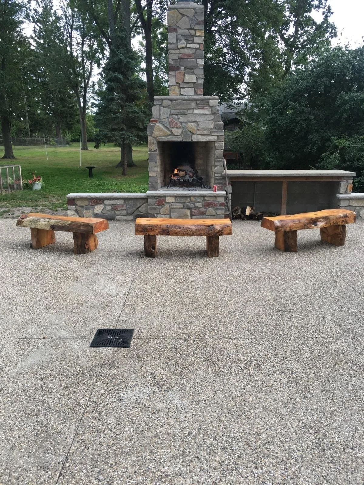 Outdoor stone fireplace with a fire burning, three rustic wooden benches in front, and a firewood storage area to the right, surrounded by trees and a gravel patio.