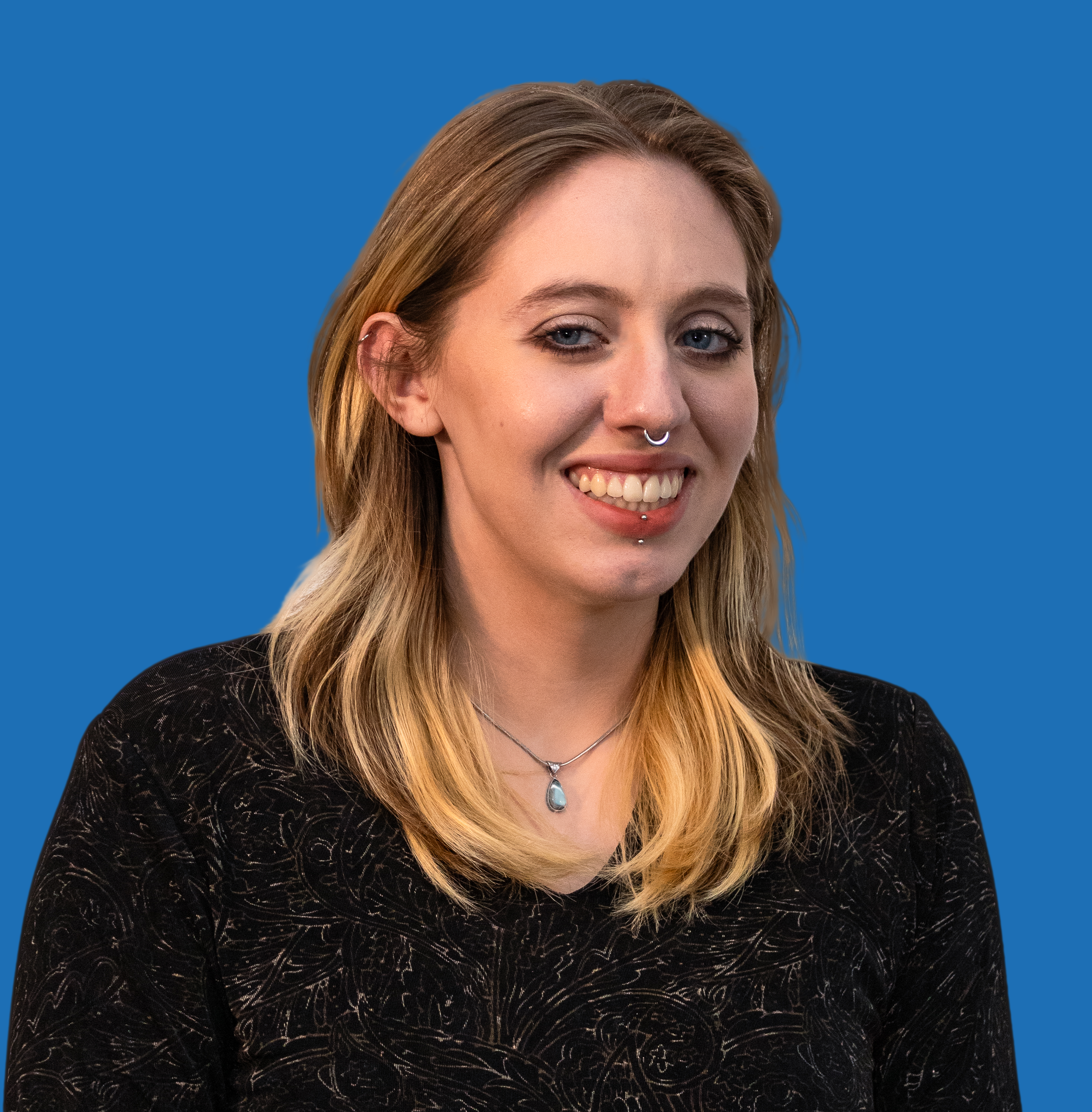 Professional headshot of website author big vibrant smile long blonde hair and glistening blue eyes. Wearing black top and medium length chain.